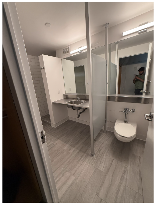 A modern lounge area with a dark ceiling featuring colorful lighting panels in blue, green, and other hues. The floor is covered in dark carpet, and two sets of gray couches with patterned upholstery are arranged around white coffee tables. To the left, a kitchenette with gray cabinets and shelves is visible through a glass wall. The space is well-lit and has a sleek, contemporary design.