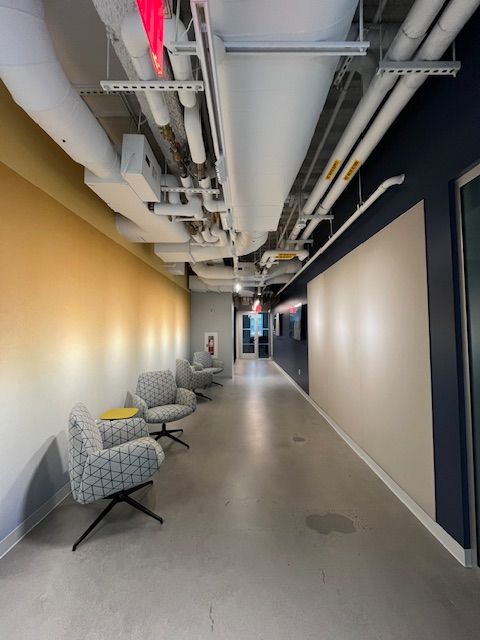 A long corridor with a smooth concrete floor. On the left side, the wall is painted in a gradient from gold to white, while the right side is blue. Two modern, patterned chairs are positioned against the left wall, one with a yellow object on it. Various pipes and ventilation ducts run along the ceiling, and a door at the far end of the hallway leads outside.
