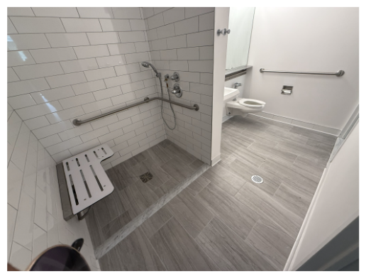 A spacious accessible bathroom with a large shower area featuring white subway tile walls, a handheld showerhead, and grab bars. There is a fold-down bench in the shower, and a toilet adjacent to it. The floor has light gray tiles with a linear drain visible in the shower area. A mirror is mounted above the sink on the opposite wall.