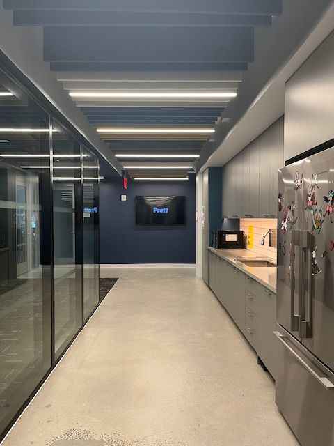 A modern office corridor features glass walls on one side, leading to a kitchen area on the right. The kitchen has light gray cabinets, a microwave, and a stainless steel refrigerator adorned with colorful magnets. A television mounted on the dark blue wall displays the word "Profit." The ceiling has decorative lighting elements, and the floor is finished in a polished concrete texture.