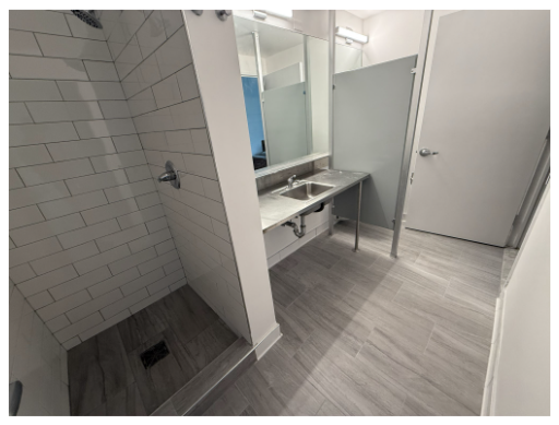 An interior view of a bathroom featuring a shower stall with gray tiled walls, a mirrored sink area with a metal countertop, and a door leading to another area. The floor is light gray and the space is well-lit, with a partially frosted glass panel for privacy.