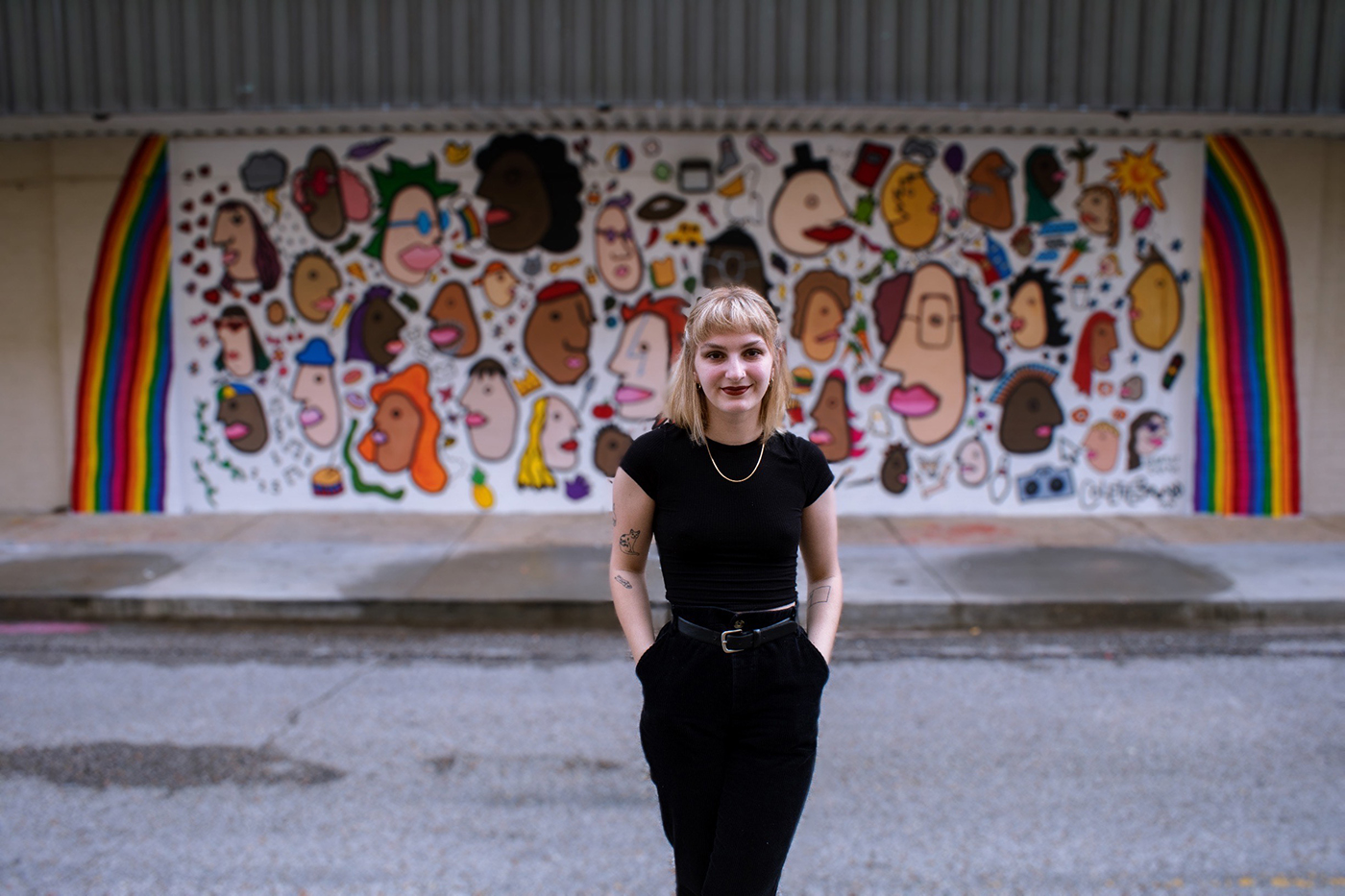 Colette Bernard stands in front of her mural I-Spy. The mural depicts diverse faces in a cartoonish style interspersed with objects.