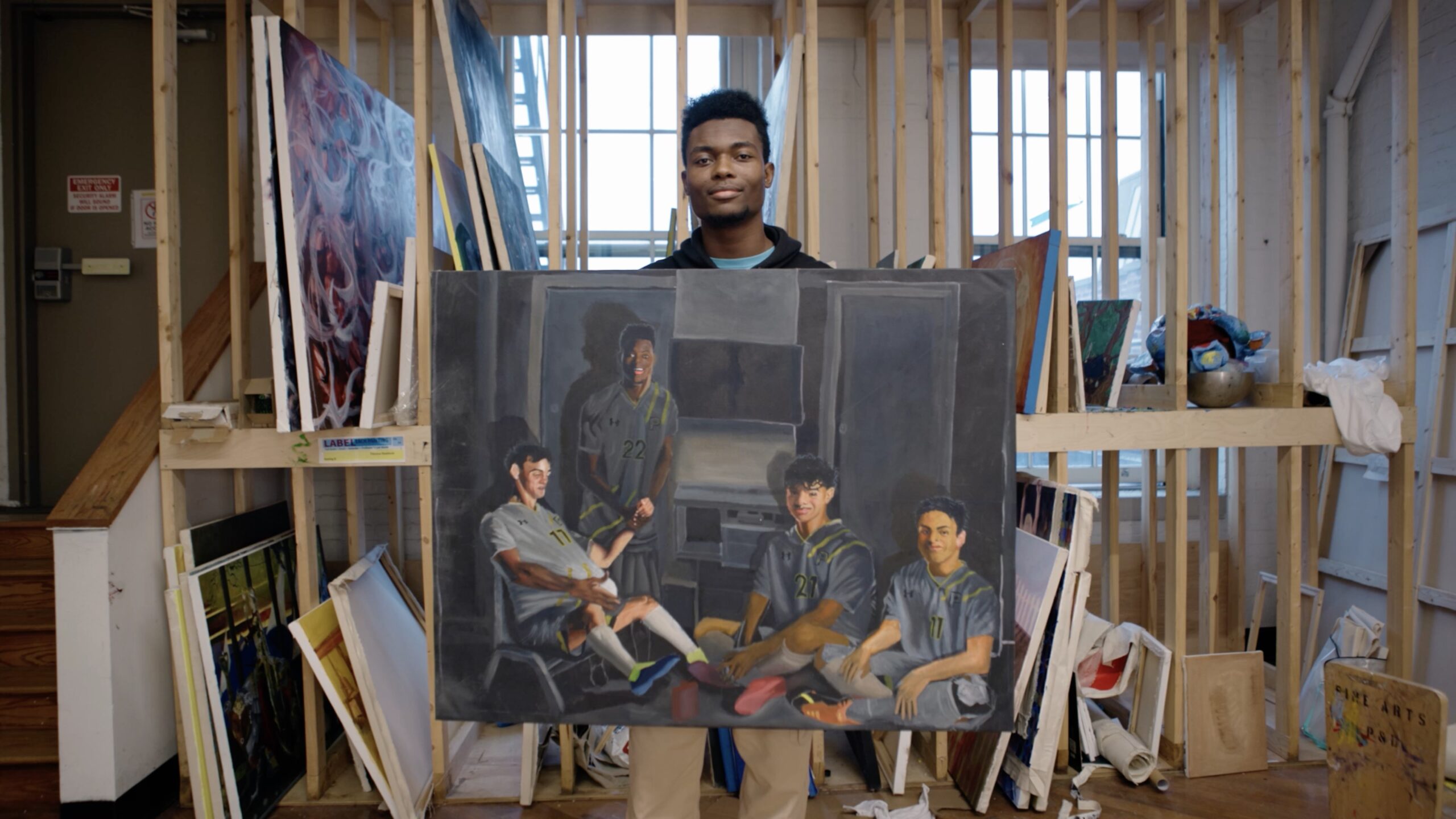 A young man stands in an art studio holding a large canvas featuring a painted scene of four young men in gray sports uniforms, seated and interacting in a relaxed setting. The background reveals various artworks and supplies, while windows let in natural light. The atmosphere conveys creativity and artistic expression.