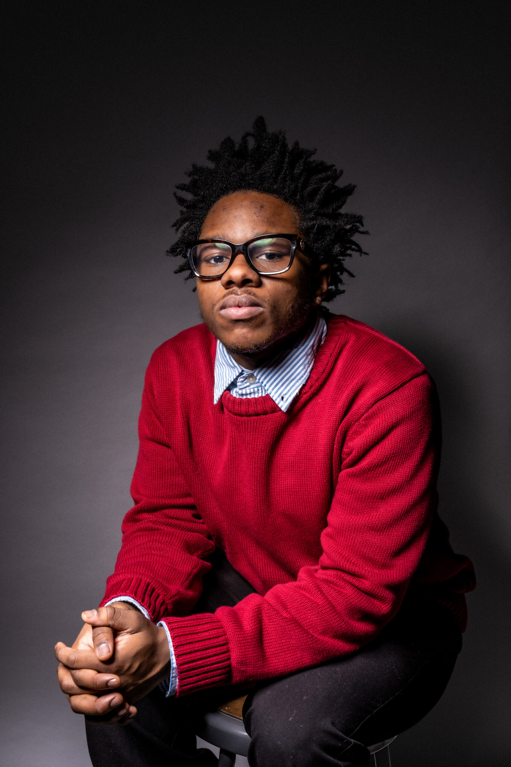 A young person with textured hair is seated on a stool, wearing glasses and a red sweater over a blue and white striped shirt. Their hands are clasped together on their knees, and they are looking directly at the camera against a dark background.