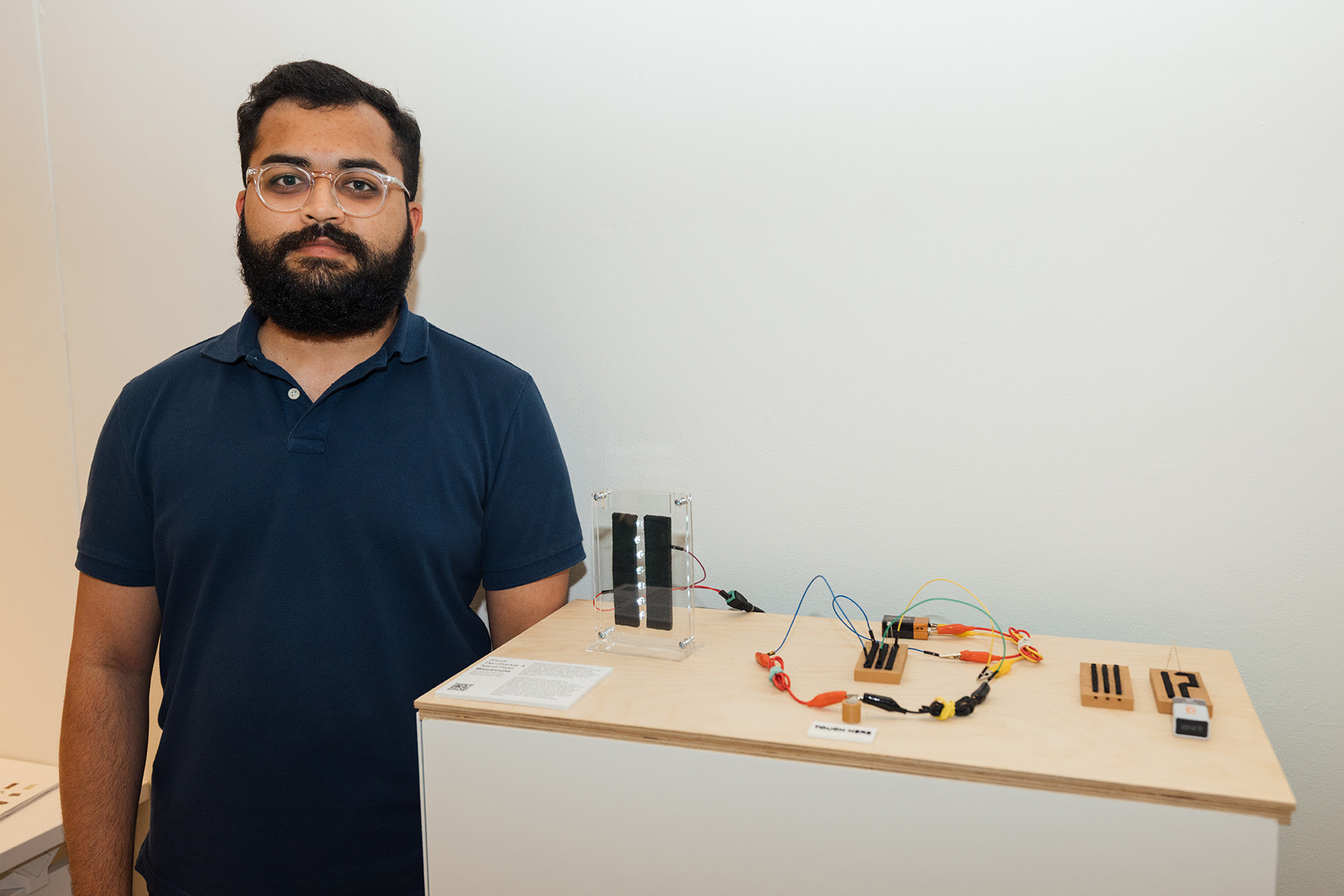 A person with a beard and glasses stands next to a display table featuring electronic components. The table has a transparent device with lights, various wires, and wooden pieces arranged on it. The background is a plain white wall, and there is printed information on the table. The individual is wearing a navy blue polo shirt.
