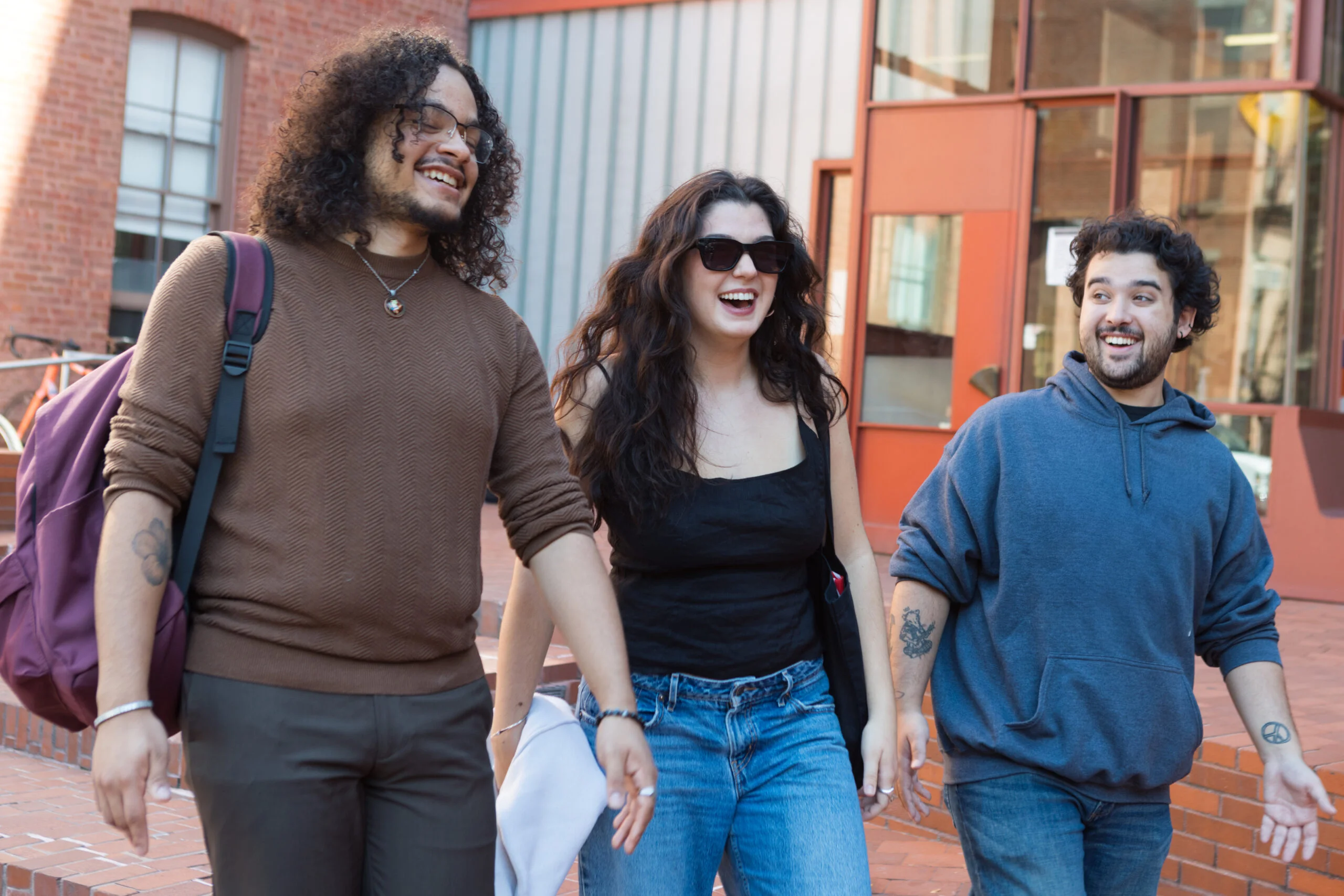 Three young adults are walking together and laughing outside. The person on the left has curly hair and a brown sweater with a backpack. The woman in the center has long, wavy hair and is wearing sunglasses and a black top, while the person on the right is in a blue hoodie and has short curly hair. They are in a brick setting with a glimpse of a building behind them.