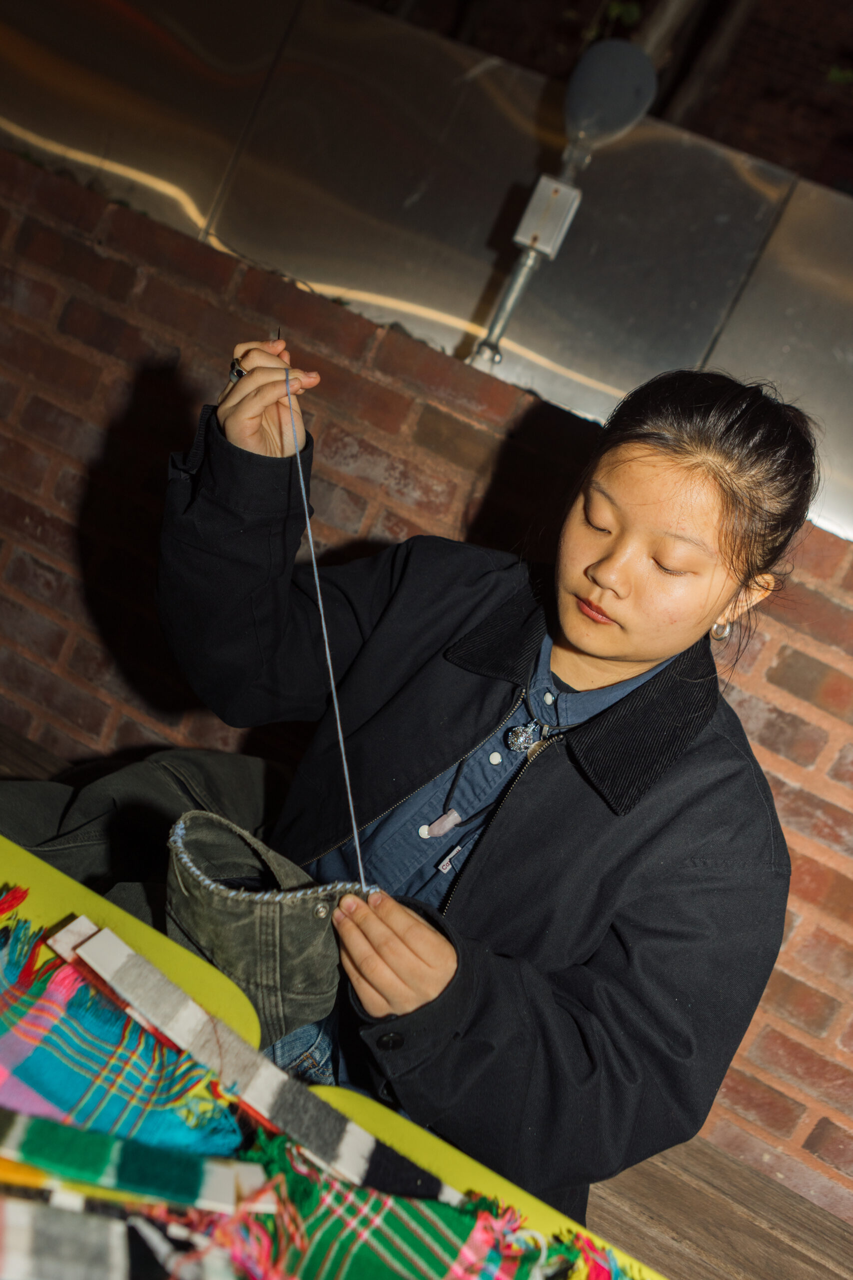 A person is sitting at a table, meticulously threading a needle with a piece of string. They are wearing a dark jacket over a blue shirt and have long hair pulled back. Various colorful fabric pieces are spread out on the table, and a brick wall can be seen in the background. The setting appears to be indoors with soft lighting.
