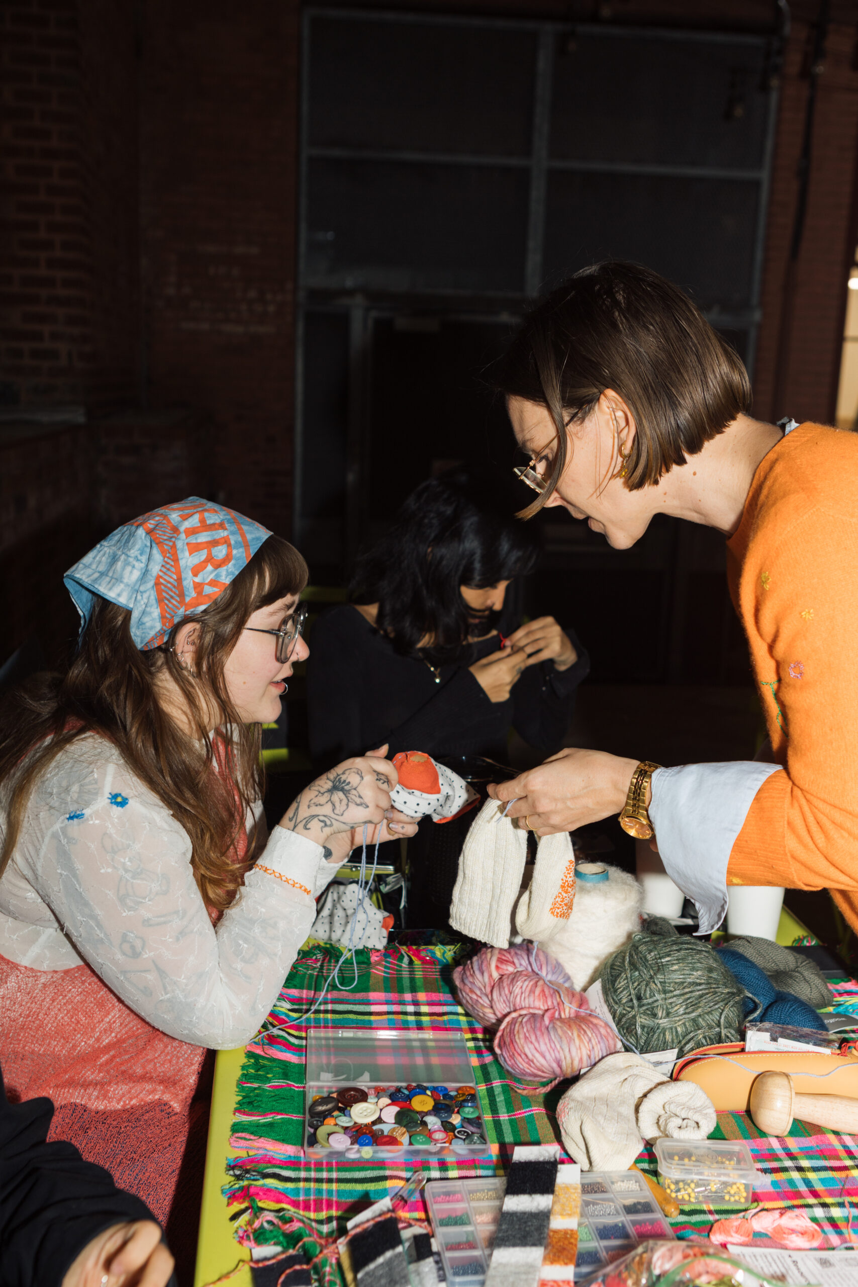 Two people are engaged in a crafting activity at a table covered with various supplies. One person, wearing a blue patterned headscarf, holds a colorful knitted piece, while the other, dressed in an orange top, hands over a piece of fabric. The table features a variety of yarns, buttons, and crafting materials against a bright plaid cloth. A third person is seen in the background, also participating in the crafting.