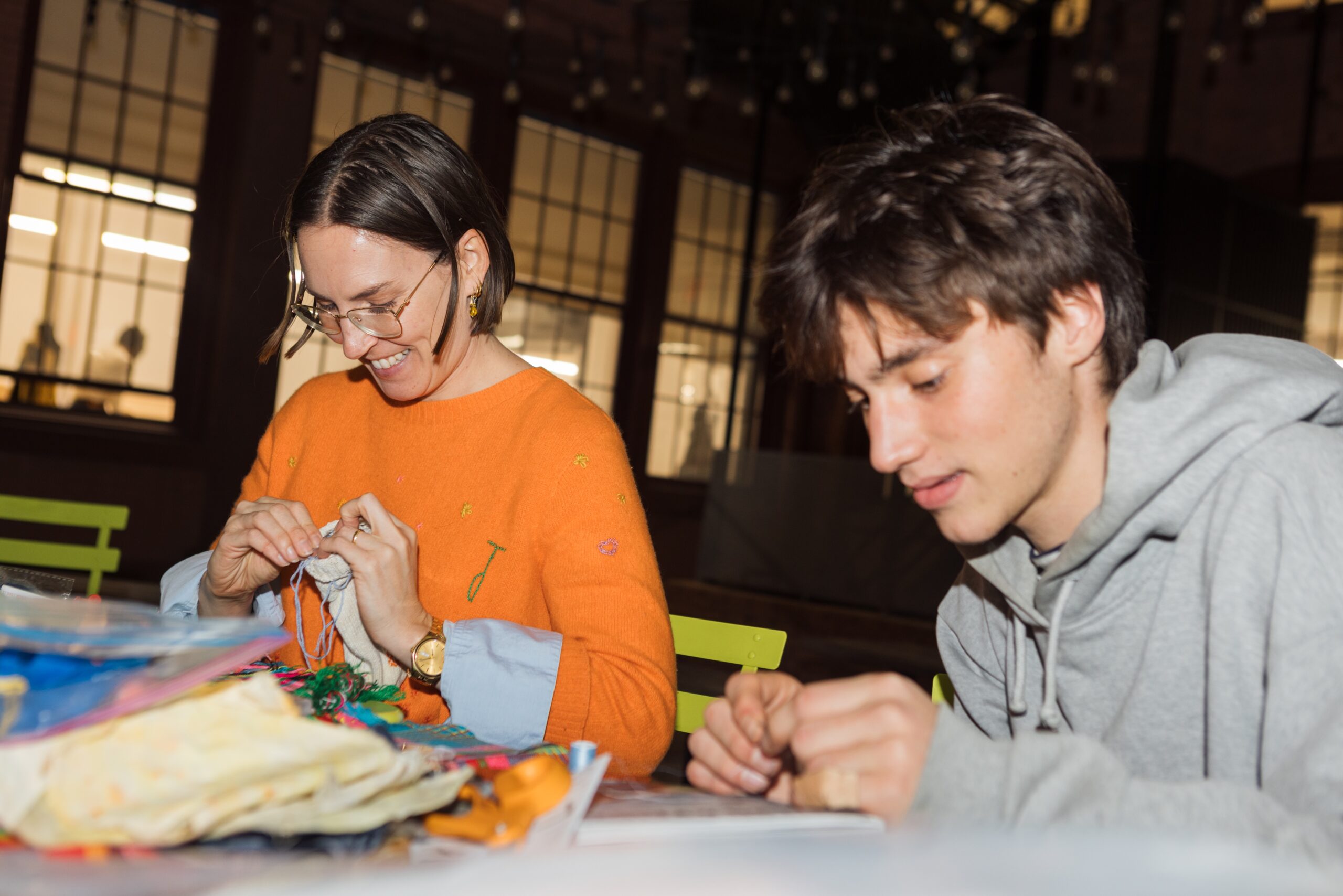 A person wearing an orange sweater and glasses smiles as she works on a craft project, while a young person in a gray hoodie focuses on a similar task nearby. They are seated at a table covered with various colorful materials and tools, in a well-lit indoor space with large windows in the background.