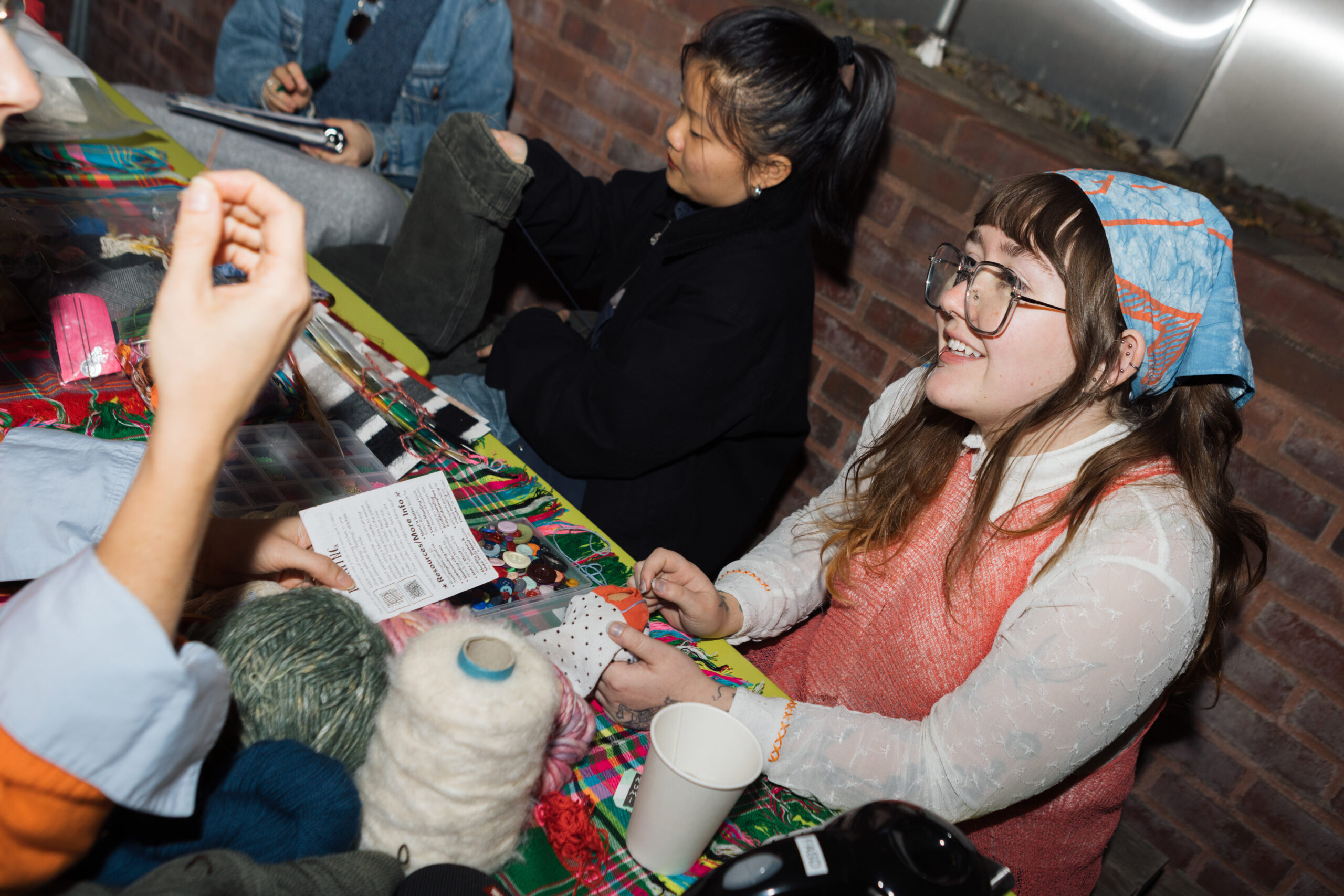 A group of people engages in a crafting activity around a brightly colored table covered with materials. One person with glasses and an orange sweater is smiling while holding a piece of fabric with a polka dot design. Another person, wearing a black jacket, is working on a project with green pants in the background. Various yarns and crafting supplies are scattered on the table.
