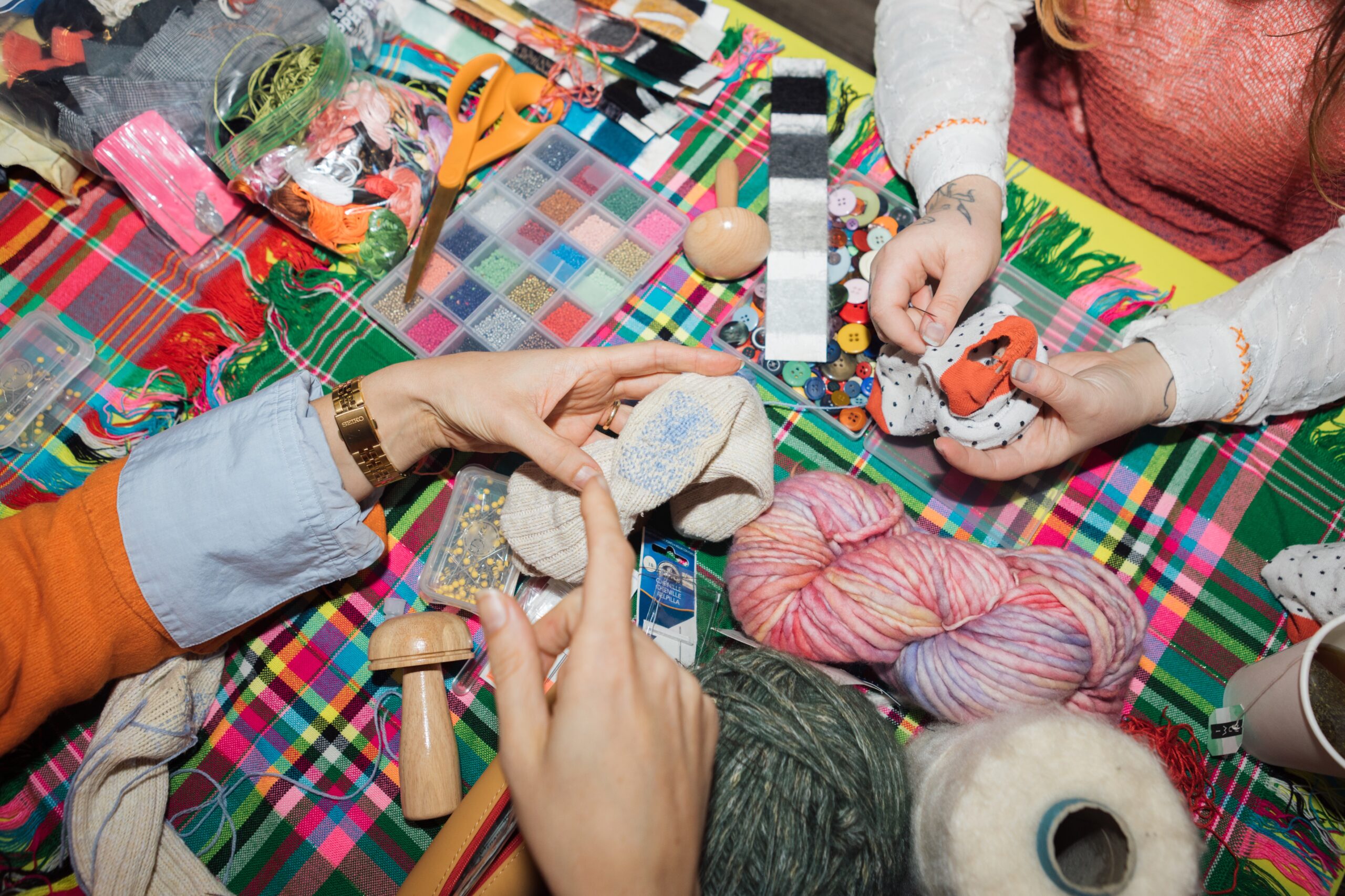 A tabletop cluttered with various crafting supplies, including colorful yarn, buttons, fabric scraps, and scissors. Two hands are visible: one holding a decorated piece of fabric, while another points towards a sock-like item with a blue pattern. A wooden tool and small containers with pins and sequins are also present on a vibrant plaid tablecloth.
