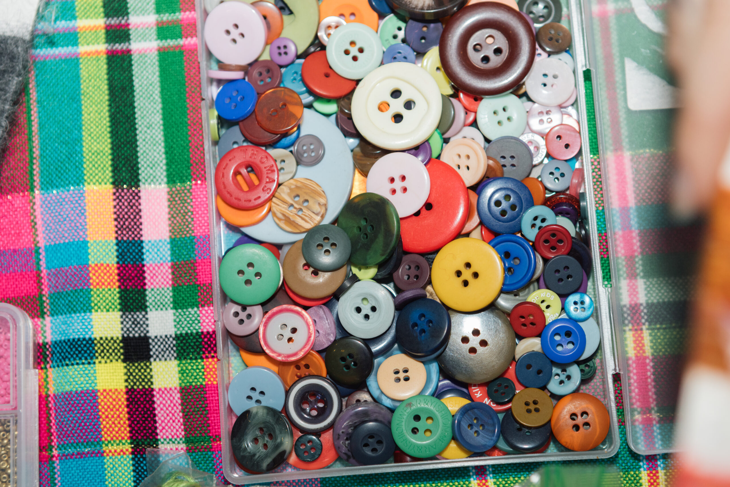 An assortment of colorful buttons in various sizes and shapes is displayed in a transparent container. The buttons feature multiple holes, with colors including red, blue, green, yellow, and orange. They are arranged atop a plaid fabric with vibrant colors like pink, green, and black, visible in the background.