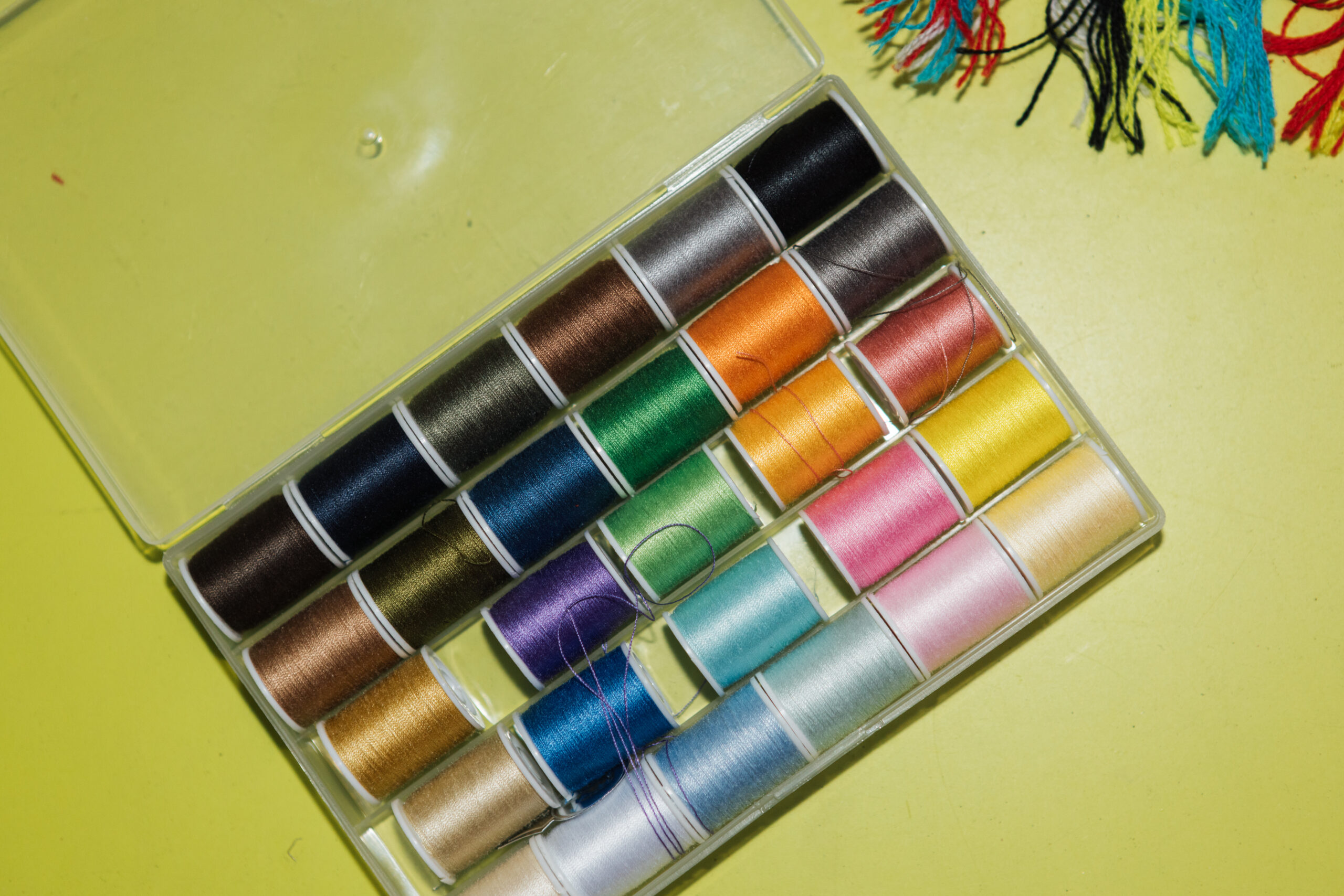 A clear plastic box containing various spools of sewing thread arranged in rows. The threads come in a variety of colors, including browns, greens, blues, yellows, and pinks. The box is placed on a bright yellow surface. In the top corner, colorful strands of embroidery floss can be seen.
