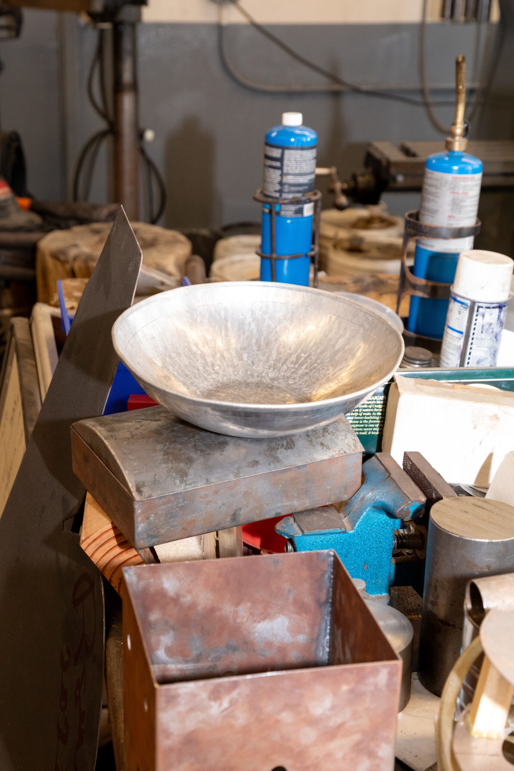 A workbench with various tools and materials. In the foreground, a shiny aluminum bowl sits atop a metal box, with a rectangular copper box nearby. Various gas canisters and a blue vice are also visible among other tools and equipment scattered across the surface.