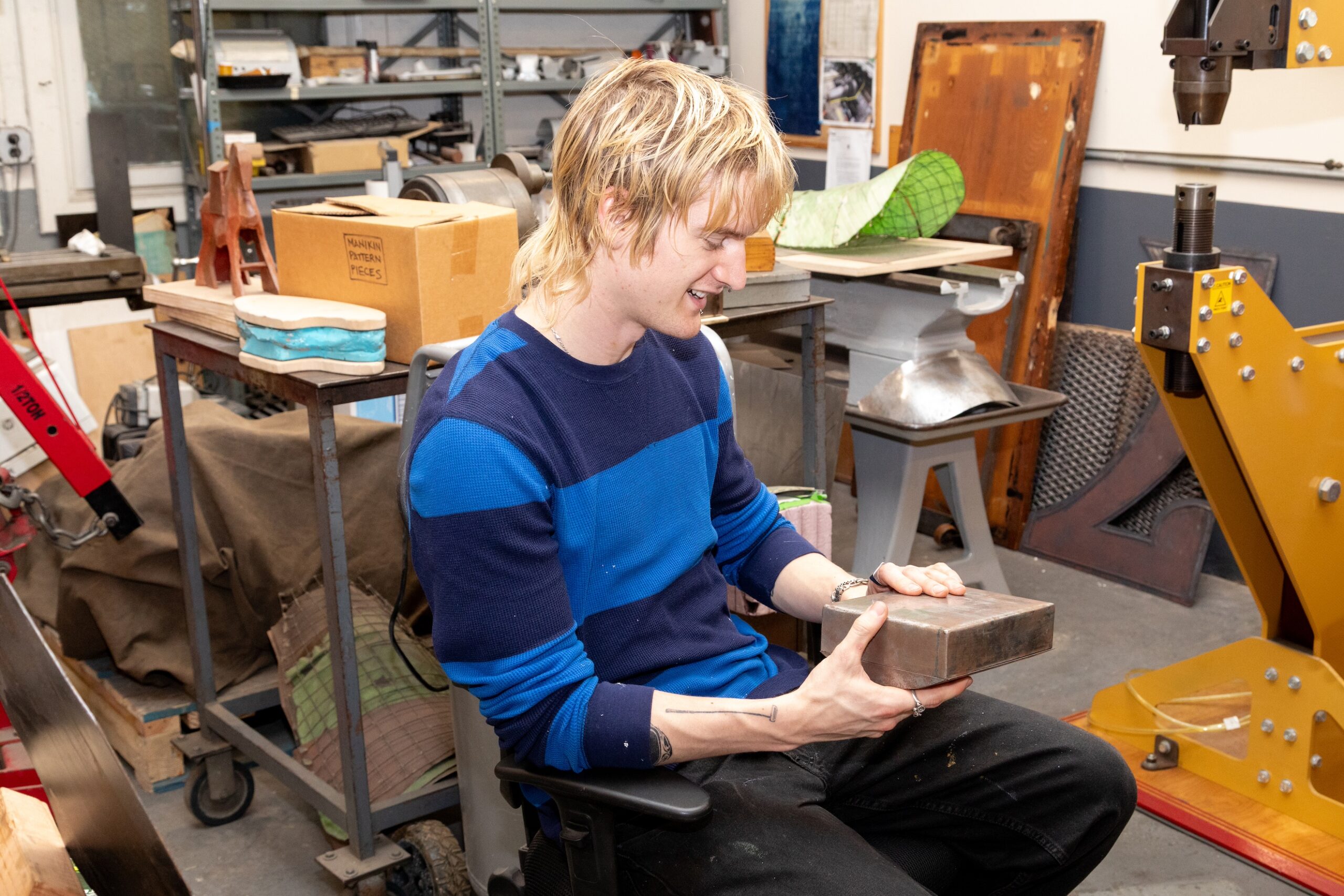 A person with blonde hair, wearing a blue striped shirt and black pants, is sitting in a workshop. They are holding a metallic rectangular block with both hands and smiling. Various tools, materials, and a yellow machine are visible in the background, an active creative workspace.