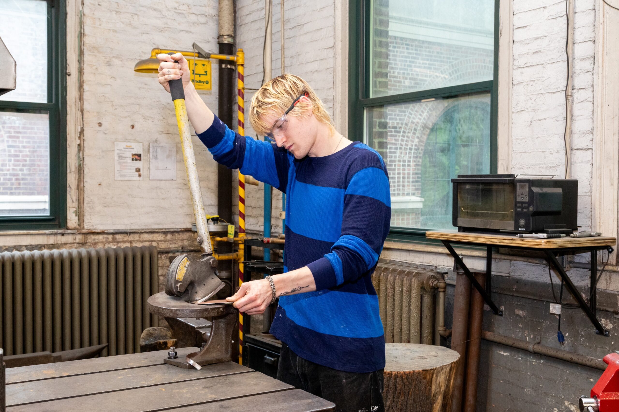 A young person with blond hair, wearing glasses and a blue and black striped shirt, is operating a machine in a workshop. They are focused on cutting a material with a press tool, positioned on a wooden workbench. In the background, there are windows, a microwave, and various workshop tools.