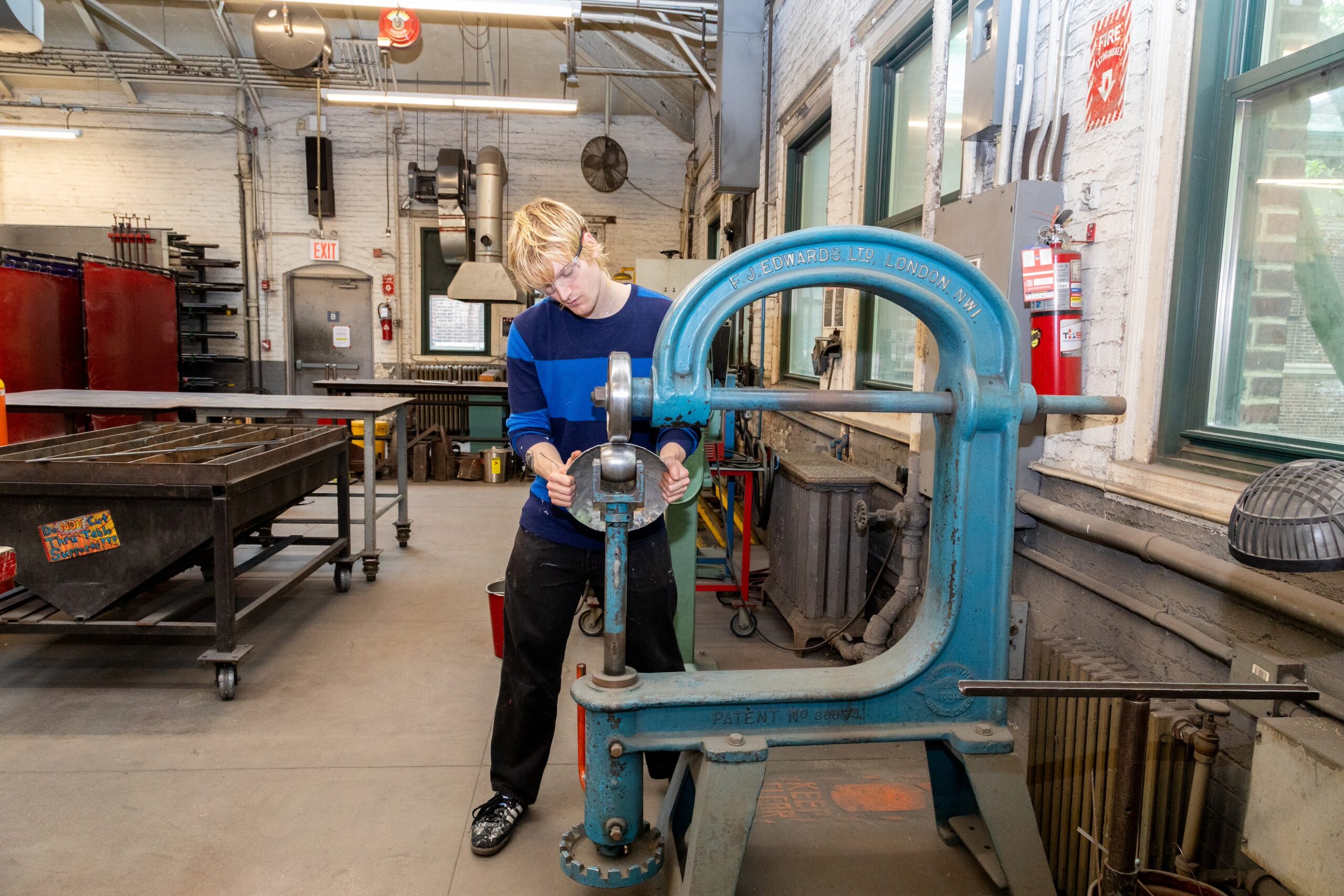 A young person with blonde hair, wearing a blue and black striped long-sleeve shirt and safety glasses, operates a large, blue metal shaping machine in a workshop. Various tools and equipment, including a metal worktable and items stored on shelves, are visible in the background, along with windows allowing natural light into the space.