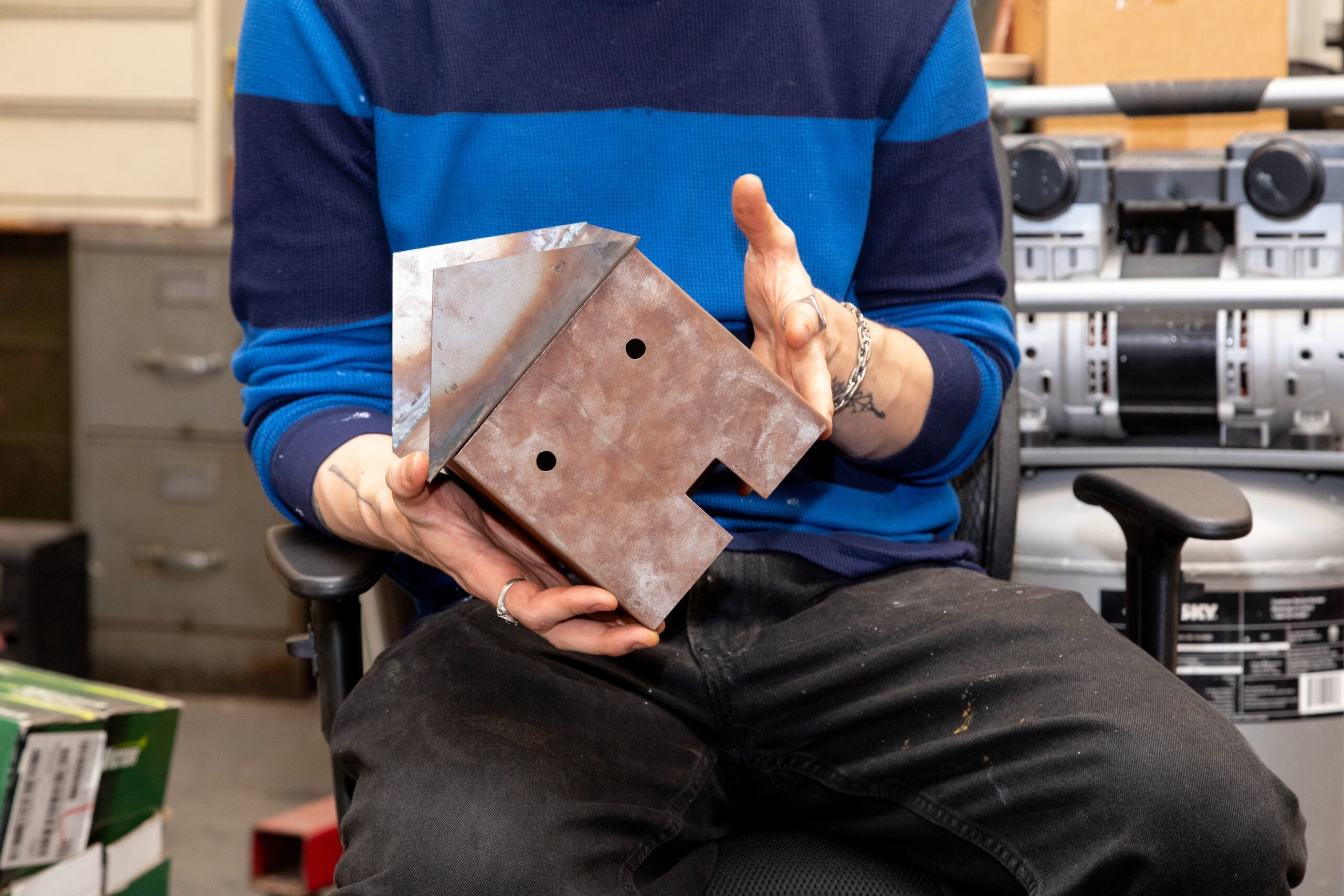 A person wearing a blue and black striped long-sleeve shirt holds a metallic object shaped like a simplified house, featuring a triangular roof and two holes in the base. The background shows storage cabinets and equipment, suggesting a workshop setting. The individual appears to be seated, with tools and materials visible nearby.