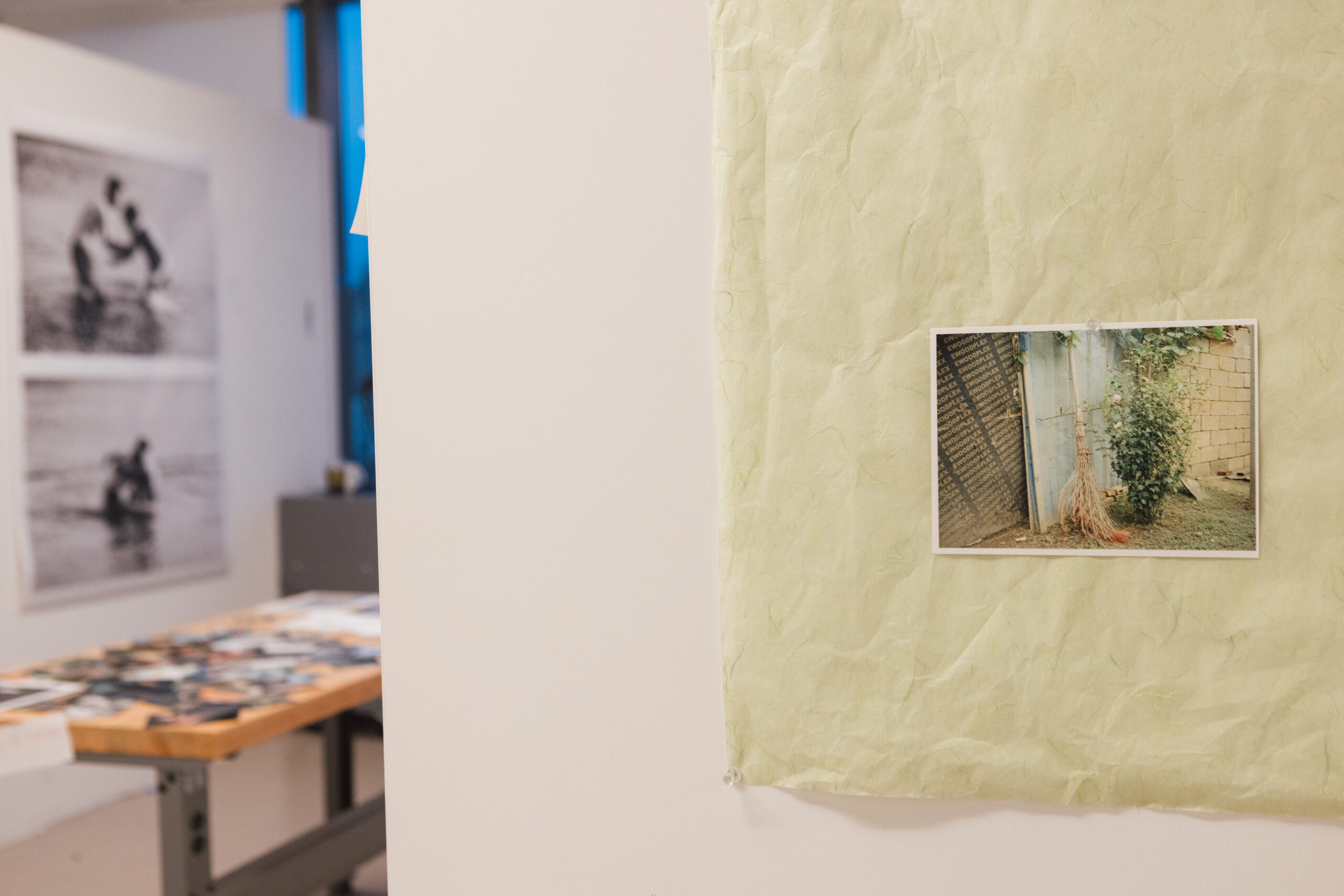 A small photograph is pinned to a textured light green sheet of paper on a white wall. The photo features a bush near a fence with a broom leaning against it. In the background, two black and white photographs are hung on another wall, depicting people in water. A wooden table with scattered images is visible in the foreground, with floor-to-ceiling windows and dim natural light.