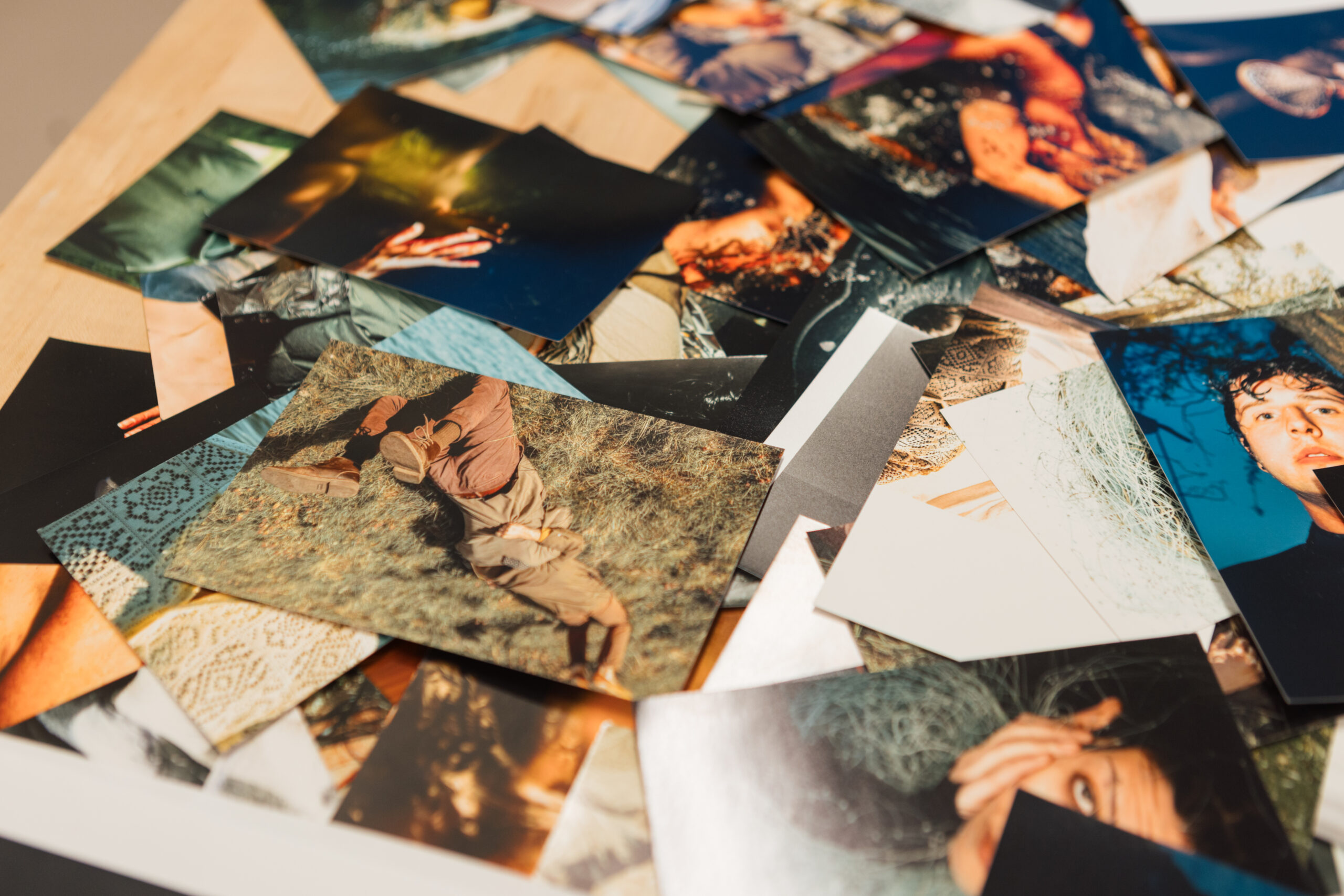 A scattered array of various photographs lies on a wooden surface. The images feature different subjects and scenes, showcasing vibrant colors and diverse compositions. Some visible photos depict a person lying on the ground, hands, and close-up facial expressions. The photographs overlap, creating a dynamic visual texture.