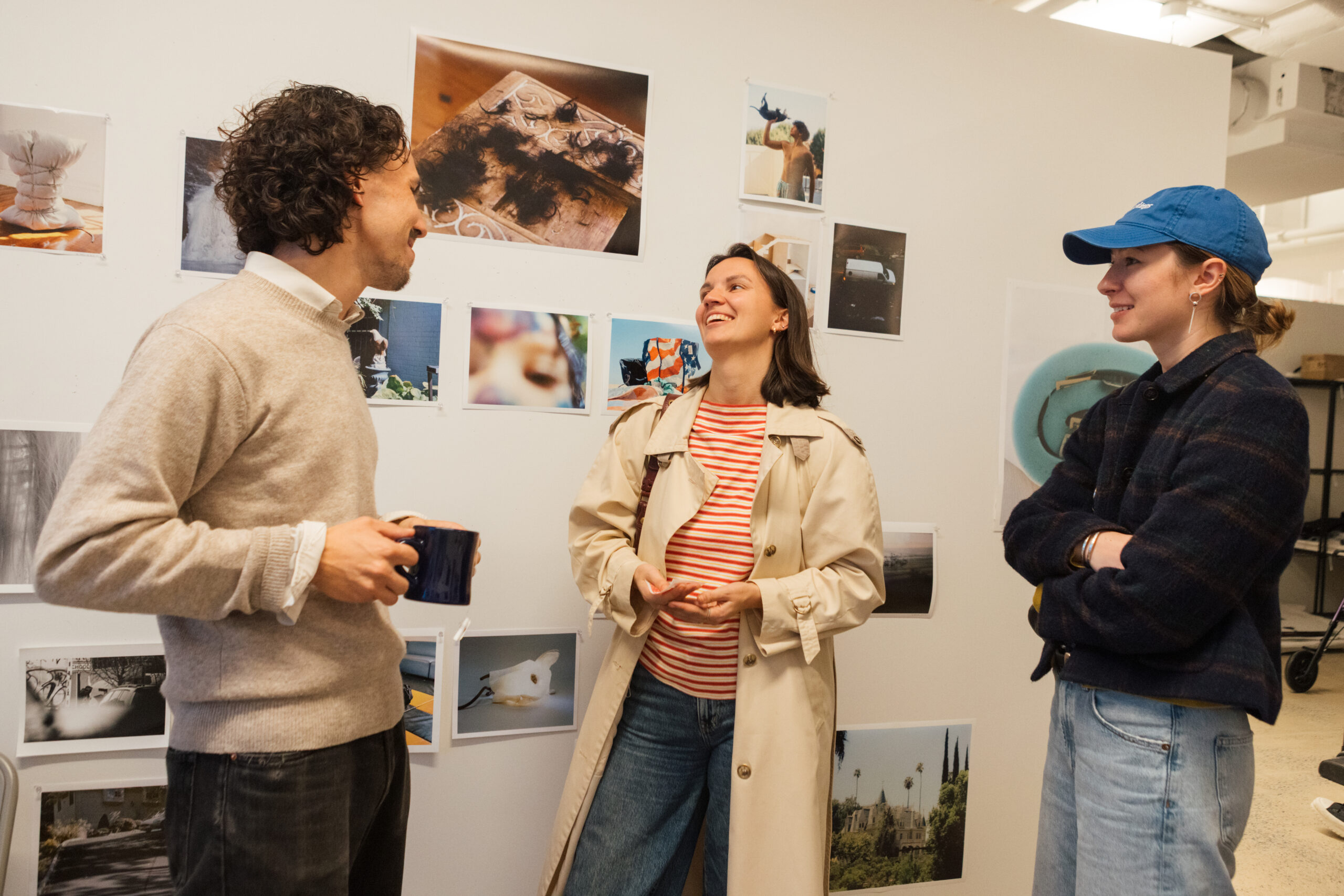 Three people are engaged in conversation in a modern space. One person is holding a blue mug, while two others are smiling; one wears a beige trench coat and striped shirt, and the other dons a blue cap and a dark jacket. Various photographs are displayed on the wall behind them, featuring different subjects and scenes.