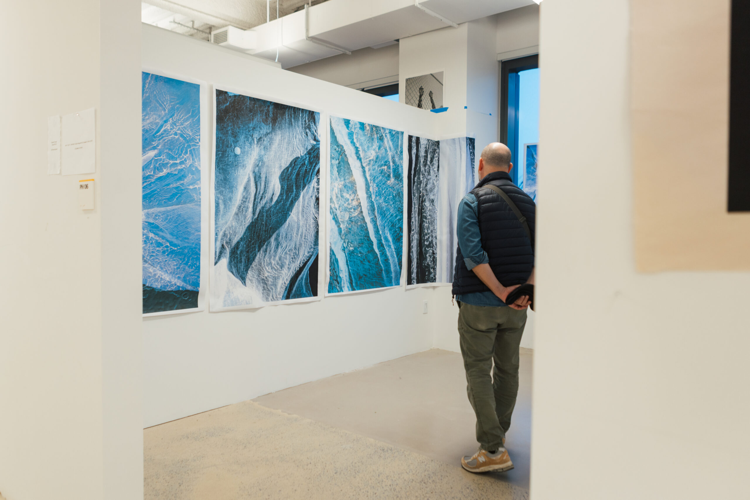 A person with short hair and a dark vest stands in an art gallery, facing a series of large, colorful photographs depicting abstract oceanic patterns in blue and black. The gallery features white walls, a concrete floor, and natural light coming through large windows. There is a piece of paper attached to the wall nearby.