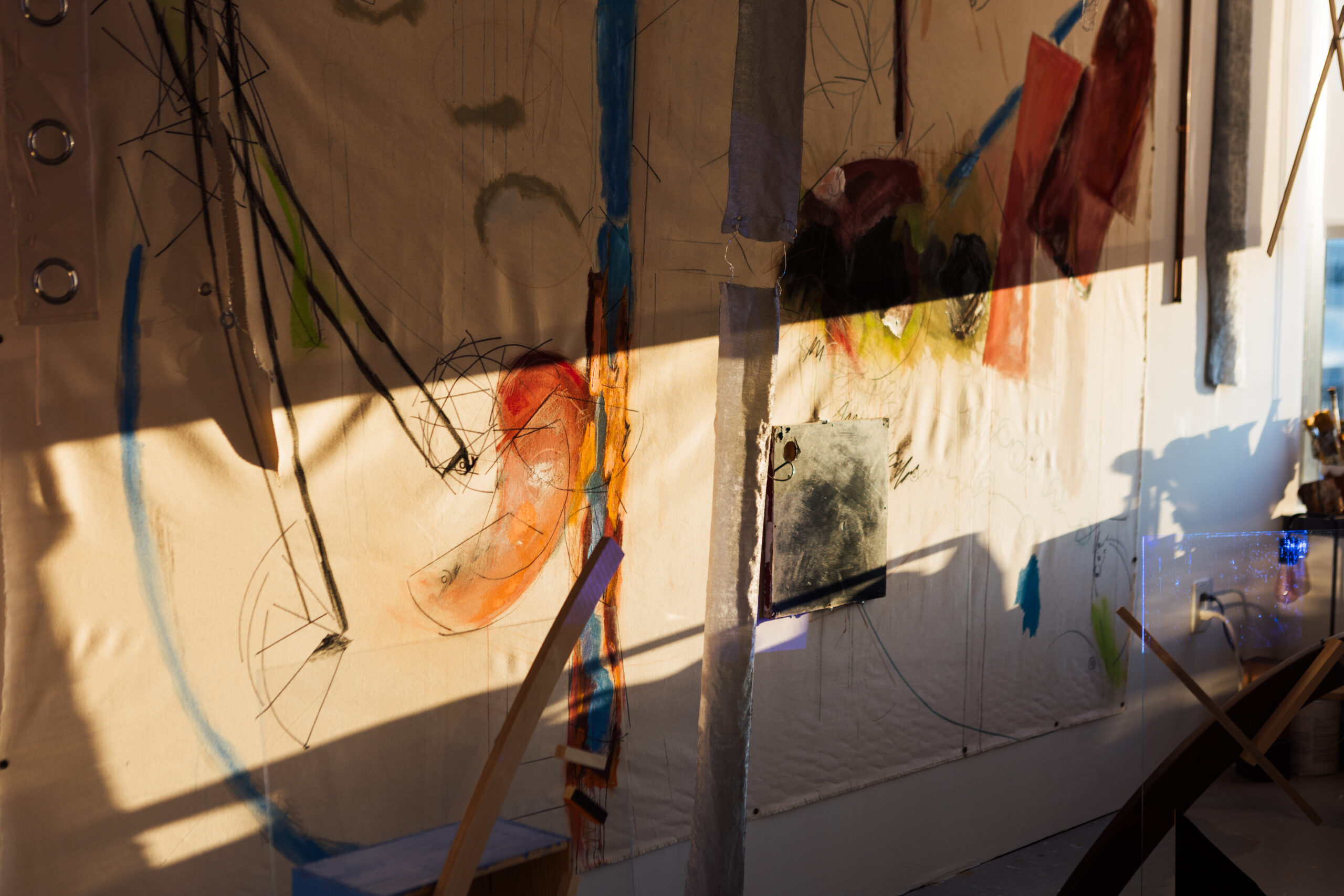 A textured wall filled with abstract artwork in various colors, including shades of red, blue, and green. The wall features sketches, lines, and circles, creating a dynamic visual effect. A shadow from an overhead structure passes across the wall, enhancing the artwork's depth. In the foreground, there's a glimpse of a wooden structure and a small table, hinting at a creative workspace. Natural light from a nearby window illuminates the scene.