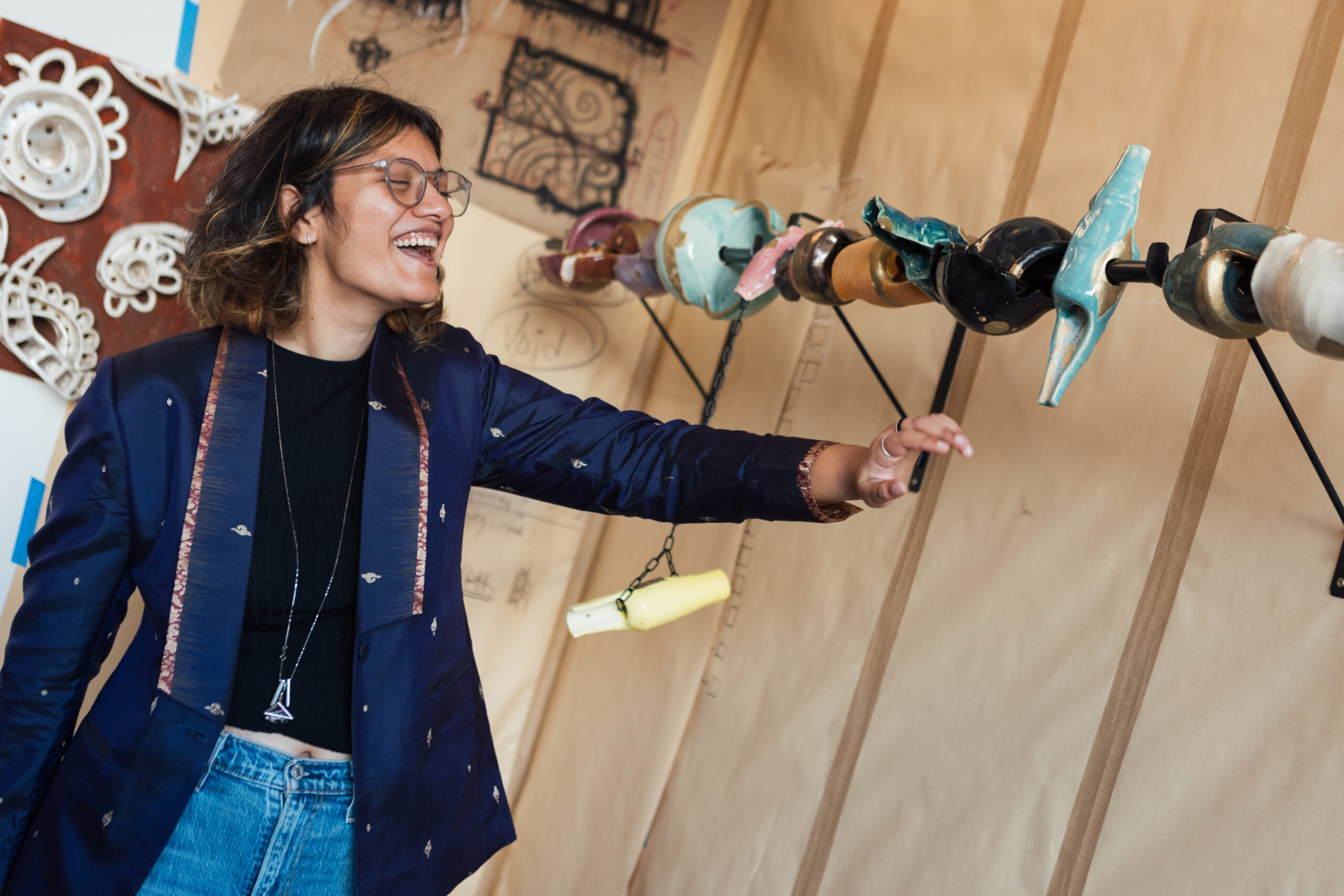 A person is smiling and reaching out towards a row of colorful objects mounted on a wall. They have shoulder-length hair, wear glasses, and are dressed in a dark blue blazer with decorative patterns and a black top, complemented by high-waisted jeans. The background features artistic designs on paper and hanging decorative elements.