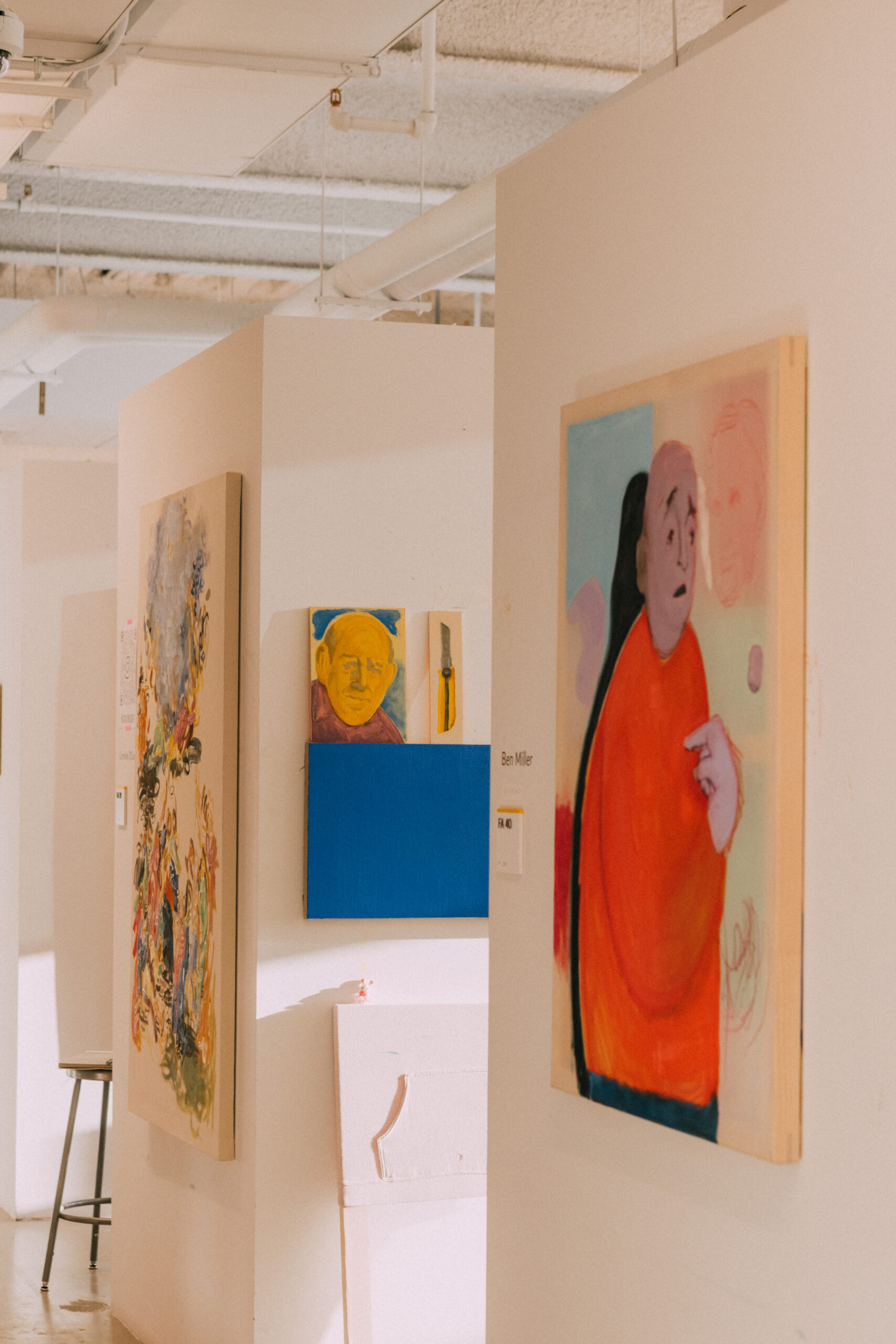 An art gallery interior featuring various artworks. To the right, a large painting of a person with an orange shirt and a concerned expression. Nearby, a blue canvas and smaller artworks, including a portrait of a man with yellow tones and a knife. The gallery has simple white walls and visible lighting fixtures, with a stool in the foreground.