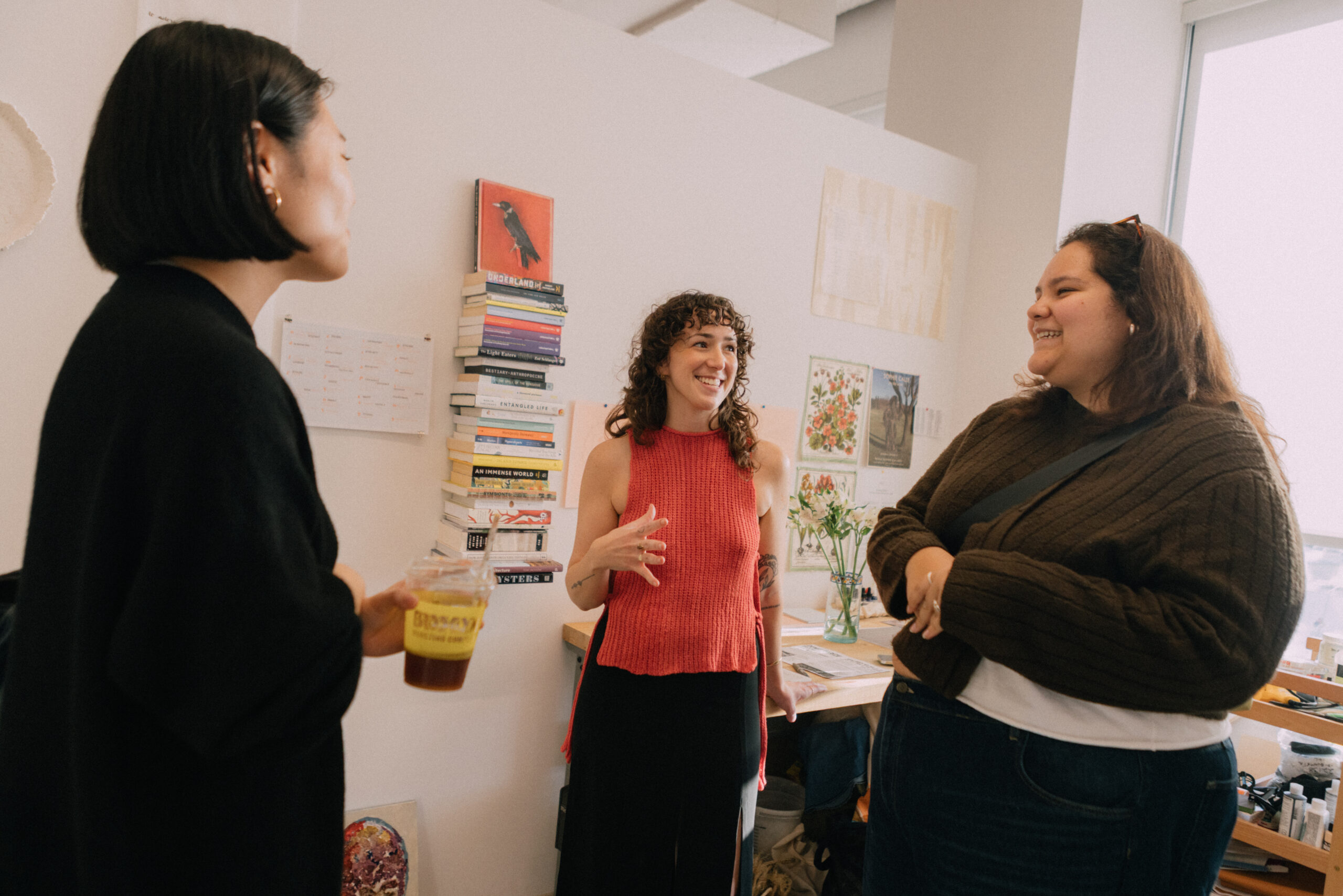 Three people are engaged in conversation in a cozy, well-lit room. One, wearing a red top, is animatedly speaking. Another, holding an iced drink, is listening attentively, while the third, dressed in a brown sweater, smiles. Behind them, there are stacked books and artwork displayed on the wall, adding to the creative atmosphere.