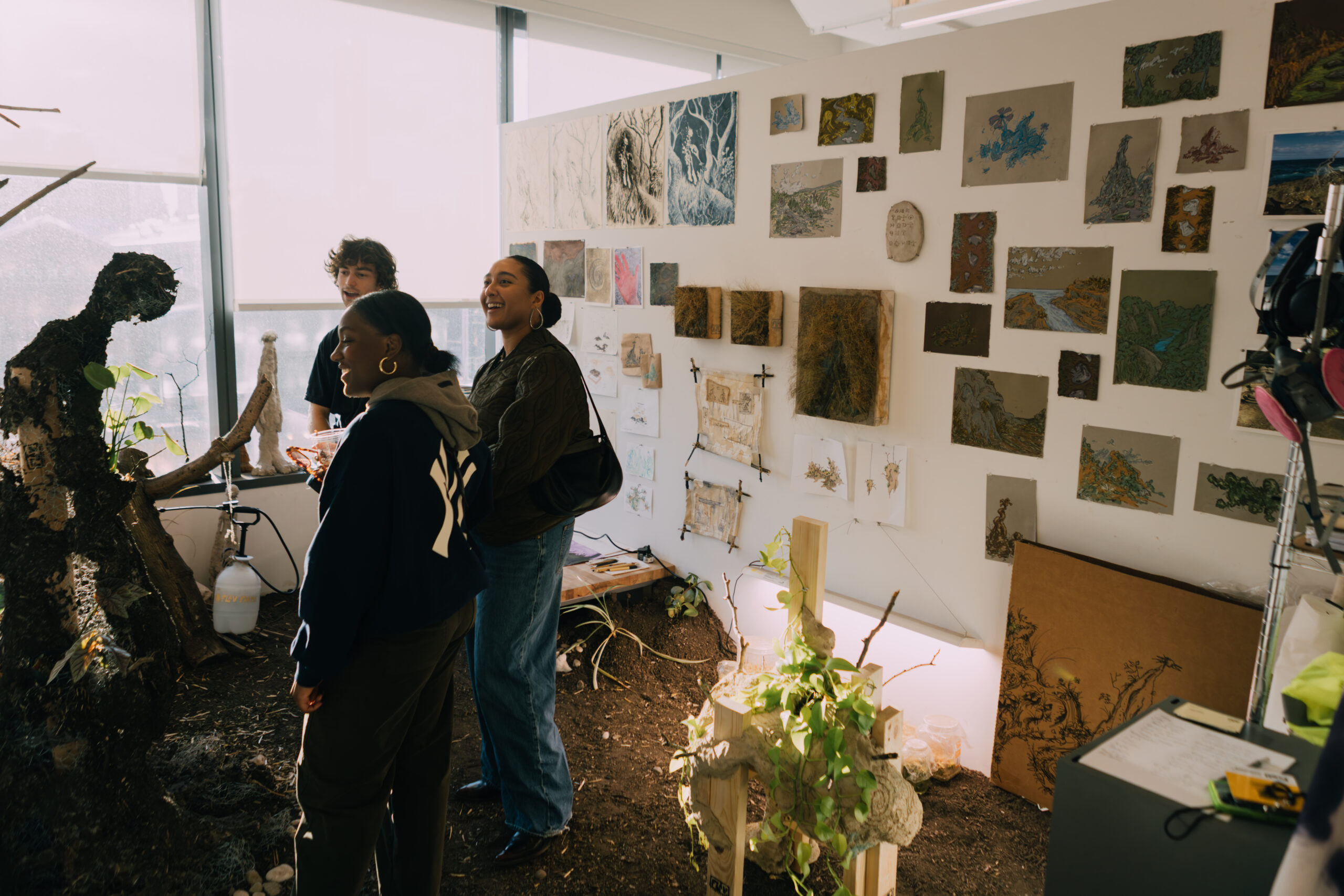Three people are indoors, observing a large art piece made from natural materials resembling a figure. The background features a wall adorned with various artistic displays, including paintings and sketches. The well-lit space includes plants, a work table, and tools on a nearby surface. The atmosphere appears creative and collaborative.