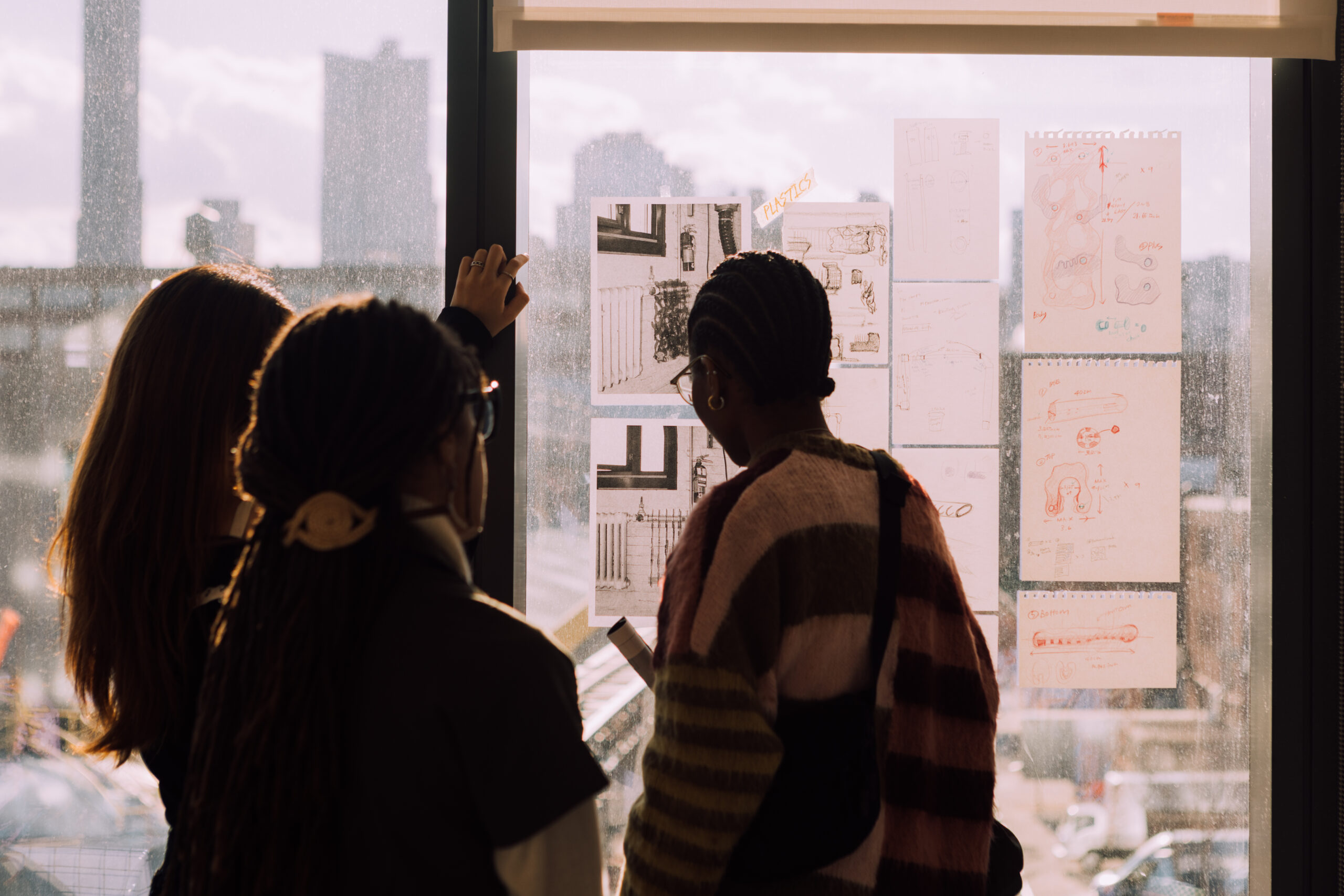 Three individuals are gathered near a window, looking at sketches and designs taped to the glass. The sunlight creates a warm glow, illuminating their shoulders and the drawings, which include various illustrations and notes. The background features a blurred cityscape of buildings.