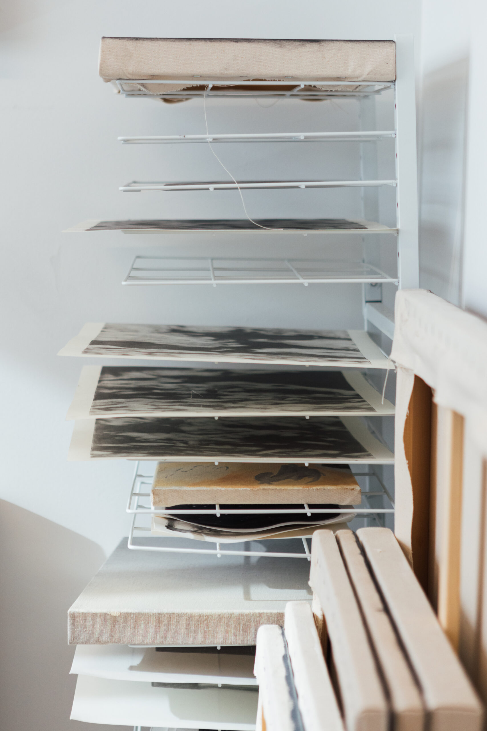 A metal shelving unit holds various pieces of artwork and paper. The top shelf is covered with a tan cloth, while several flat artworks, primarily in black and white, are arranged on the shelves below. Some pieces are partially visible, showcasing unique textures and patterns. A few canvases and padded boards are seen leaning against the side of the shelf. The background is minimal and light-colored.