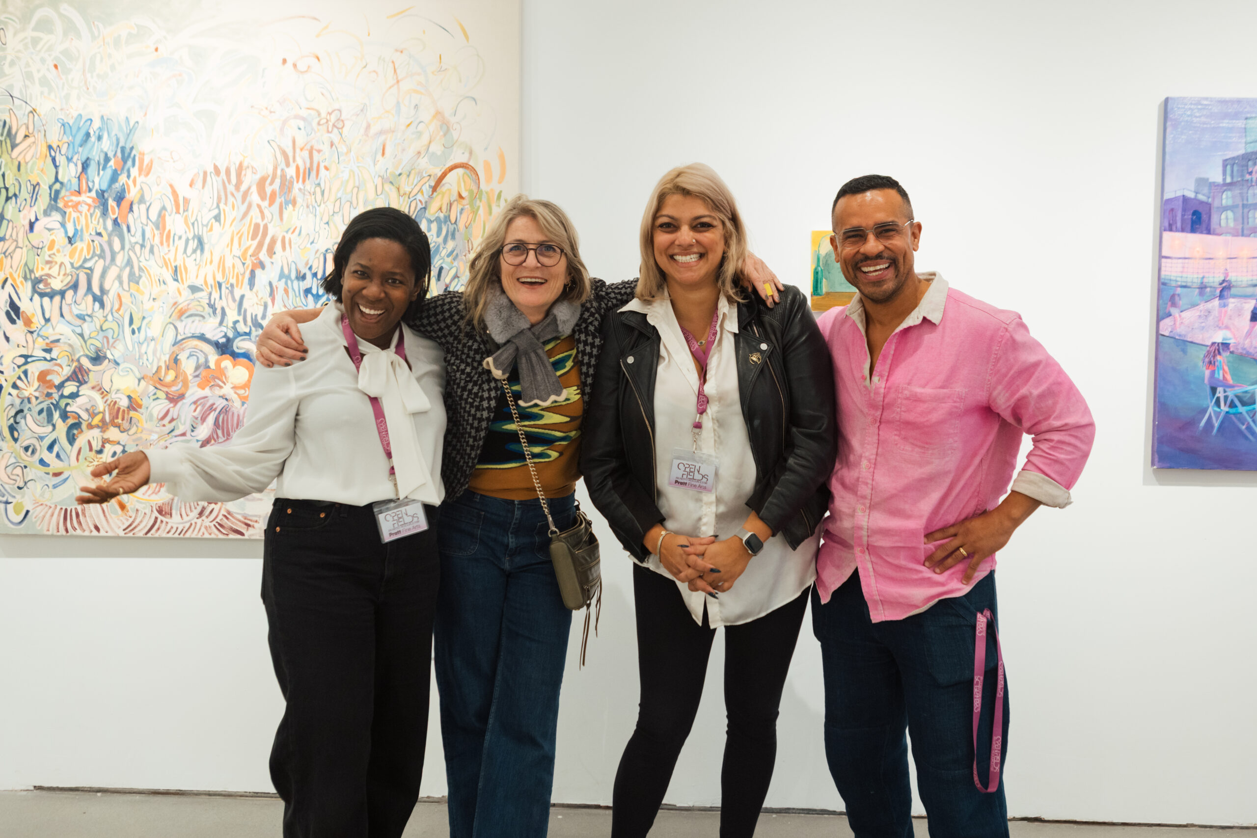 Four people stand together smiling in an art gallery, with colorful abstract paintings in the background. They appear to be enjoying their time at the event, dressed in casual attire, with name tags visible. The atmosphere feels friendly and celebratory.