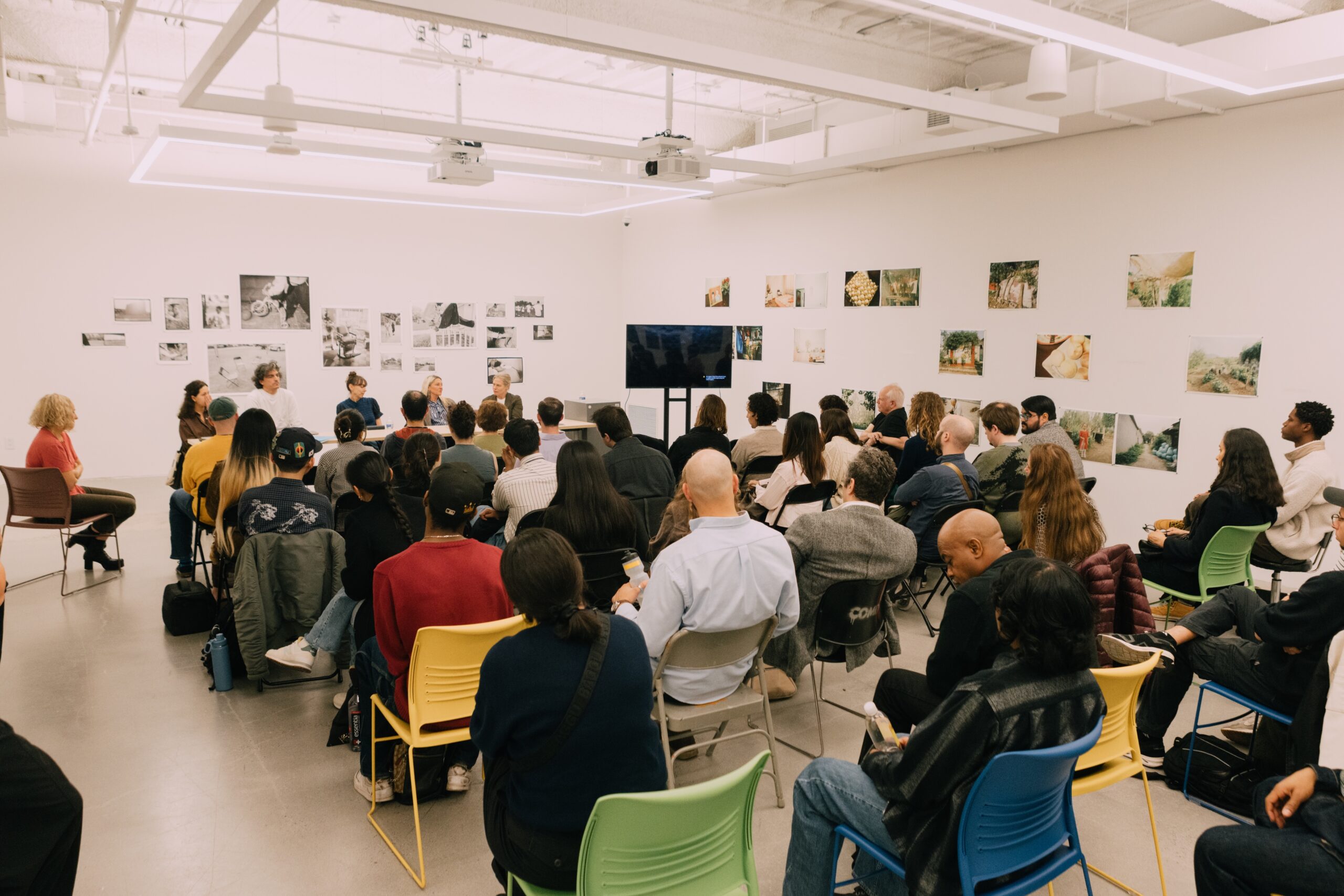 A crowded room with an audience seated on chairs, facing a panel of speakers at the front. The background features a wall adorned with various black-and-white and color photographs. A television is set up on a table near the panel, and people are engaged, some taking notes or listening attentively. The atmosphere appears to be a discussion or presentation event.