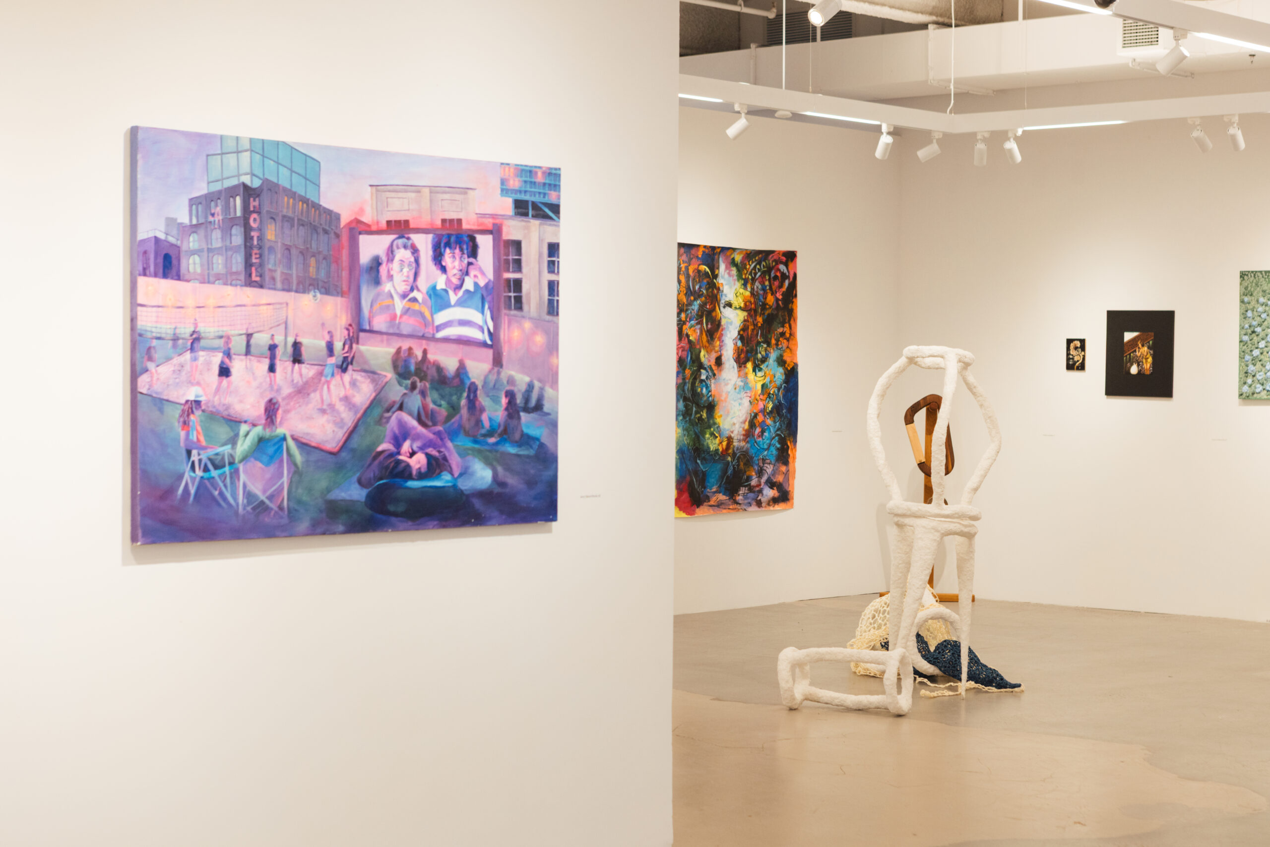 An art gallery interior features a colorful painting depicting people on a volleyball court next to a hotel. Several artworks are displayed on the walls, including a large, vibrant abstract piece, a black and white photographic work, and a unique sculpture resembling intertwined forms. The gallery has a neutral background with well-placed lighting, enhancing the visibility of the artworks.