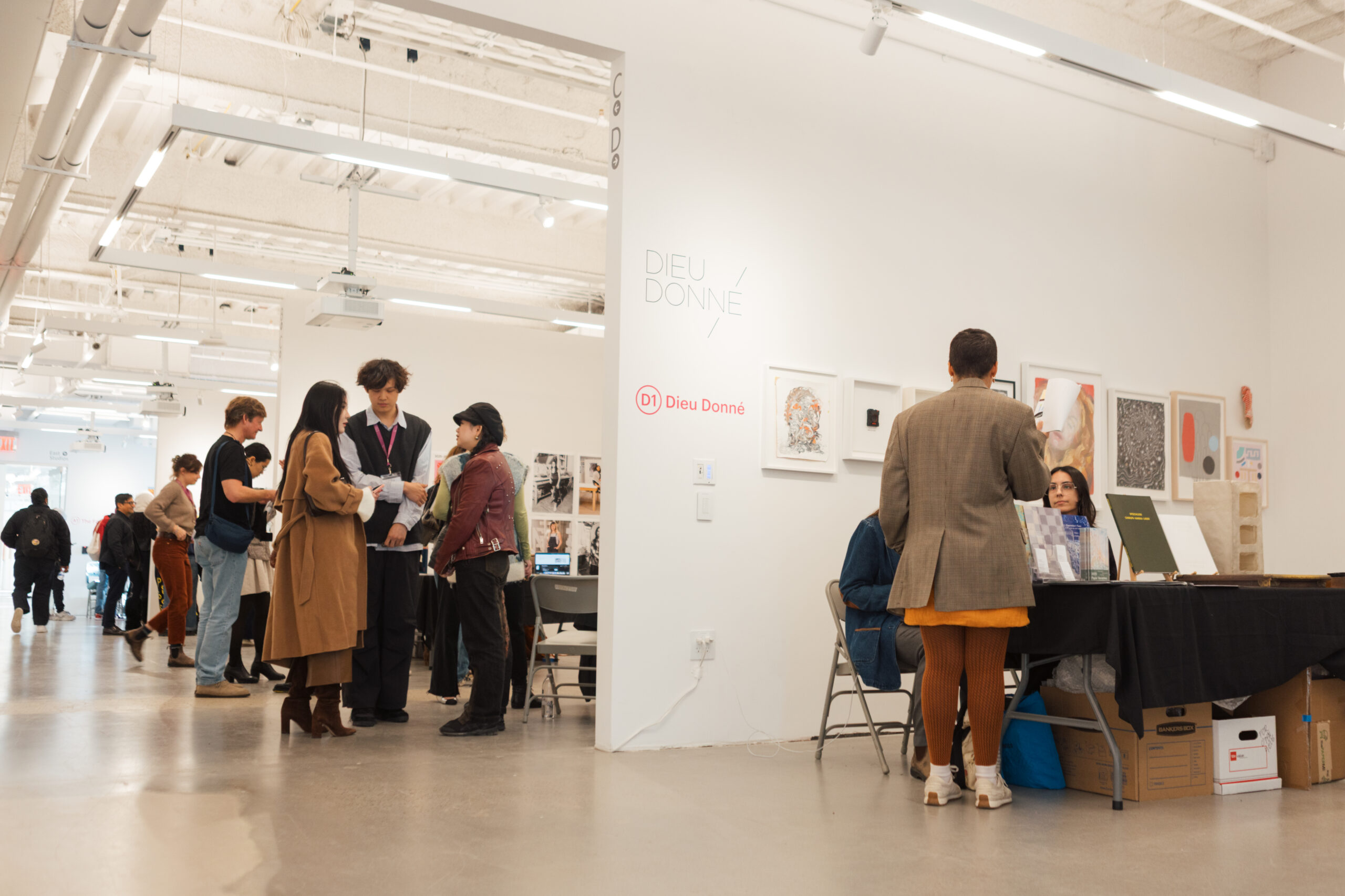 An indoor art event scene featuring Dieu Donné gallery. Several groups of people are gathered, some engaged in discussions while others browse artworks displayed on walls and tables. A person in a brown coat and boots talks to individuals in front of a table with various art materials and publications. The space is well-lit with modern decor, showcasing art pieces framed on the wall.
