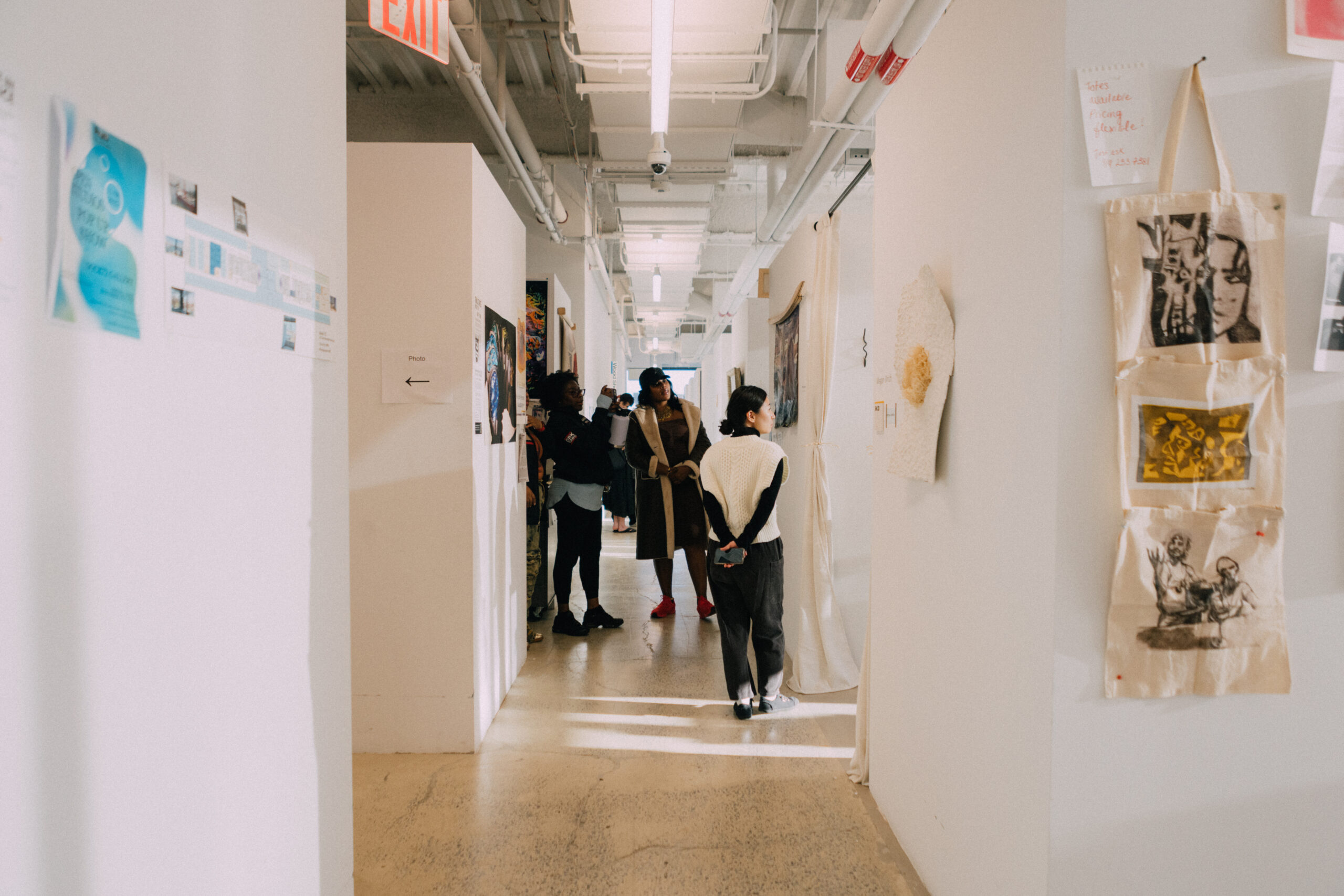 A spacious indoor hallway with white walls displays various artworks. Several people are gathered, including a person in a knitted sweater looking at an artwork. Other individuals stand nearby, engaging in conversation. Art pieces and printed materials are pinned or hung along the walls, contributing to an engaging atmosphere. An exit sign is visible above, indicating the direction to the nearest exit.