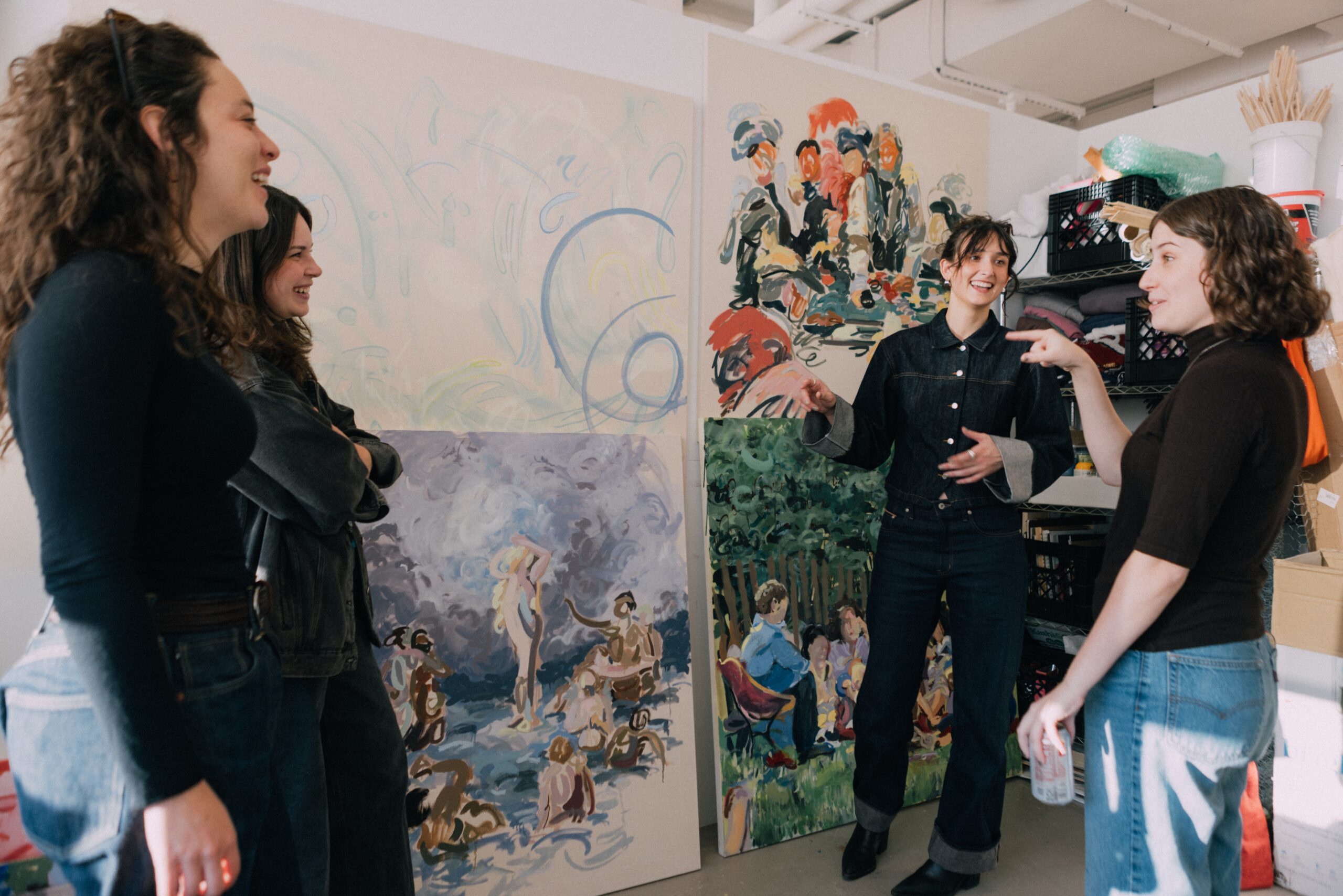 Four people are engaged in conversation in an art studio surrounded by large canvases. Two people are laughing, while another gestures animatedly. The artwork behind them features colorful and expressive scenes, depicting figures in various settings. The environment is bright and creative, with art supplies visible in the background.