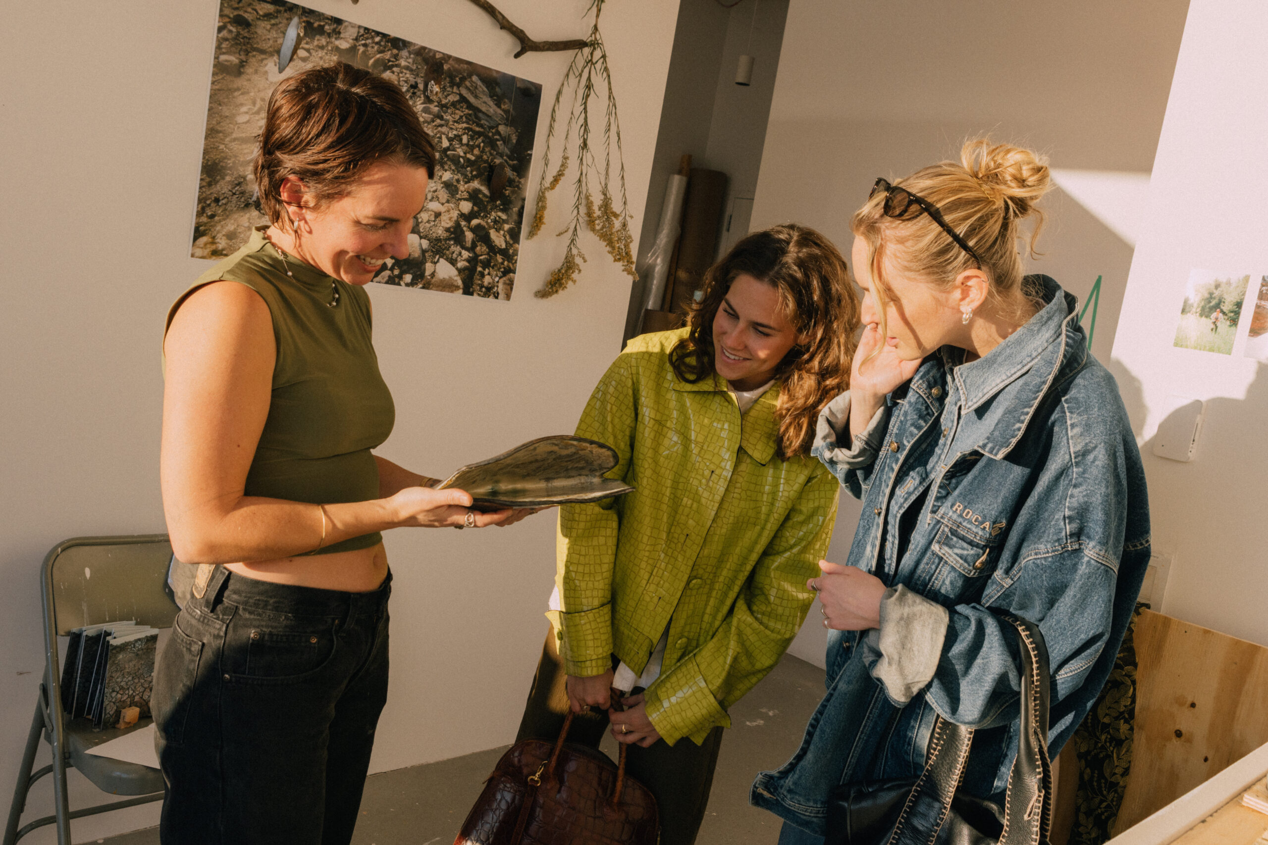 Three people are engaging in conversation inside a brightly lit room. One person, wearing a green crop top and black pants, is showing a book to the others. The second person, dressed in a bright green jacket, smiles as they look at the book. The third person, in a denim jacket, appears amused and is holding their hand to their face. The background features artwork and decor elements on the walls.