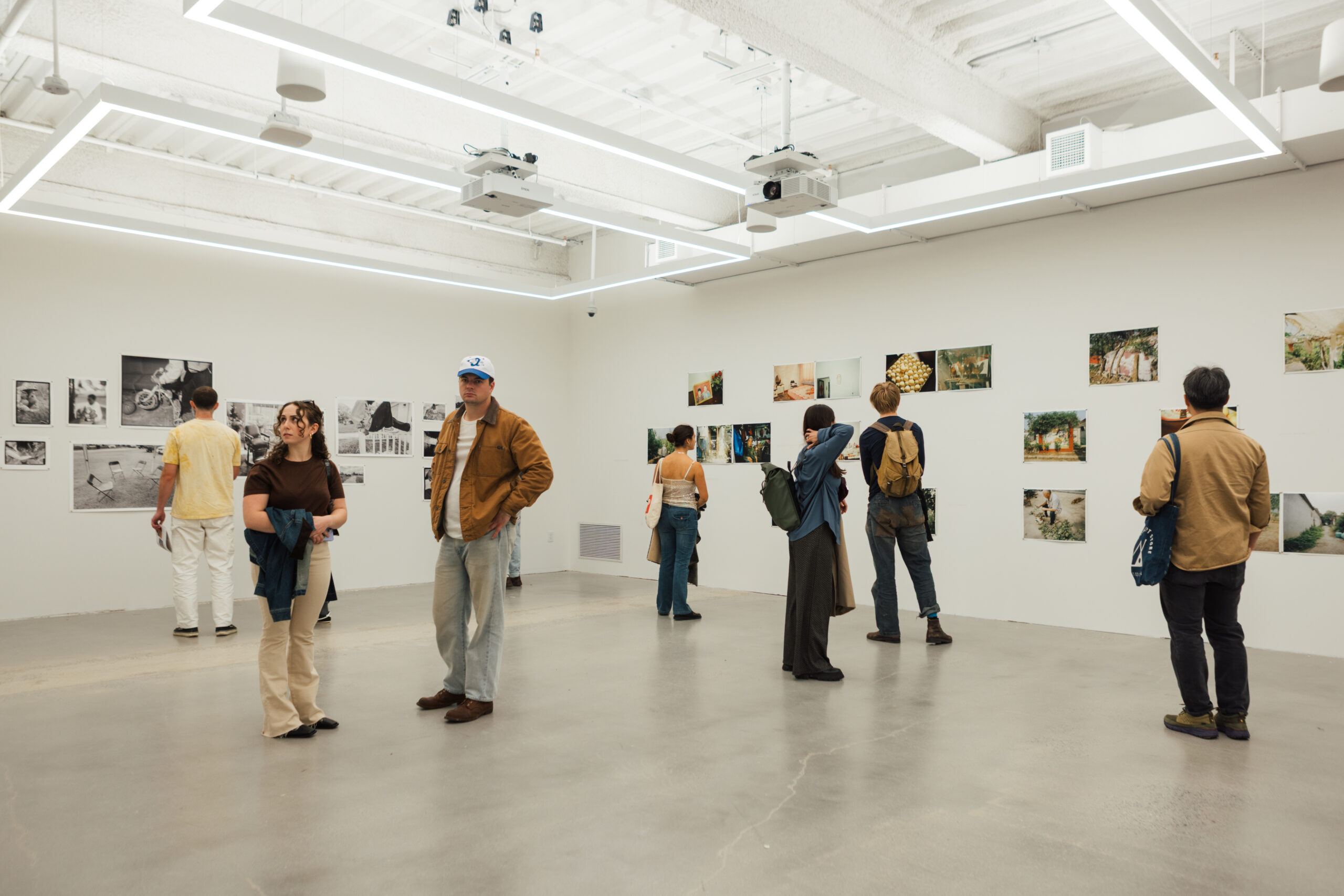 Visitors explore an art gallery featuring several photographs displayed on white walls. Four people are engaged with the exhibits, while others are positioned throughout the space. The interior is well-lit with modern ceiling lights, and the floor is a light-colored concrete.