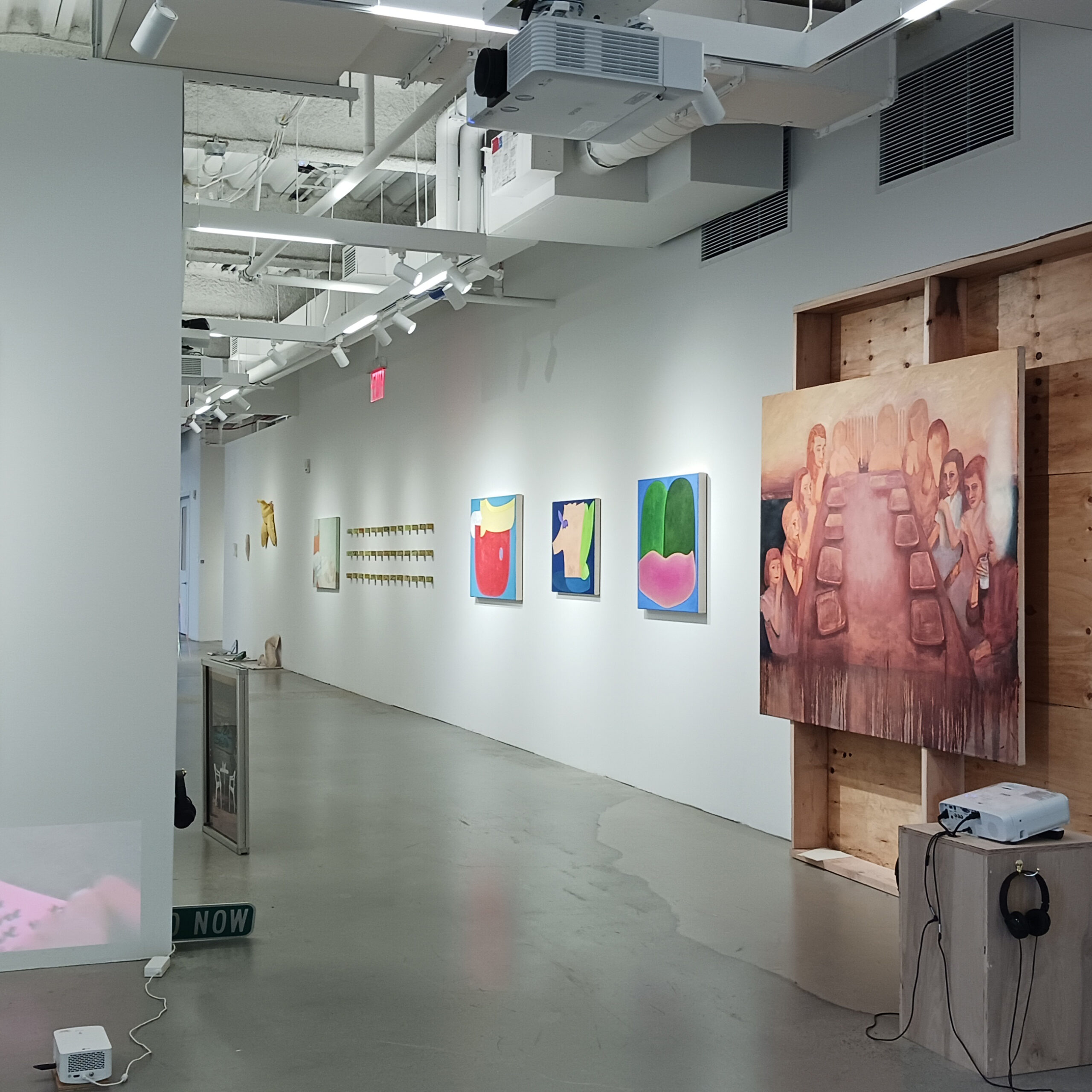 Installation view of Spring 2025 MFA Thesis Show 2 at Dock 72 in the Brooklyn Navy Yard. A long wall going down a hall has a painting installed over a wooden frame structure. In the immediate foreground are two projects facing opposite walls. In the subsequent bay, partially blocked by the wall, is a street sign and upright-standing framed image. On the long wall are installations of multiple paintings.