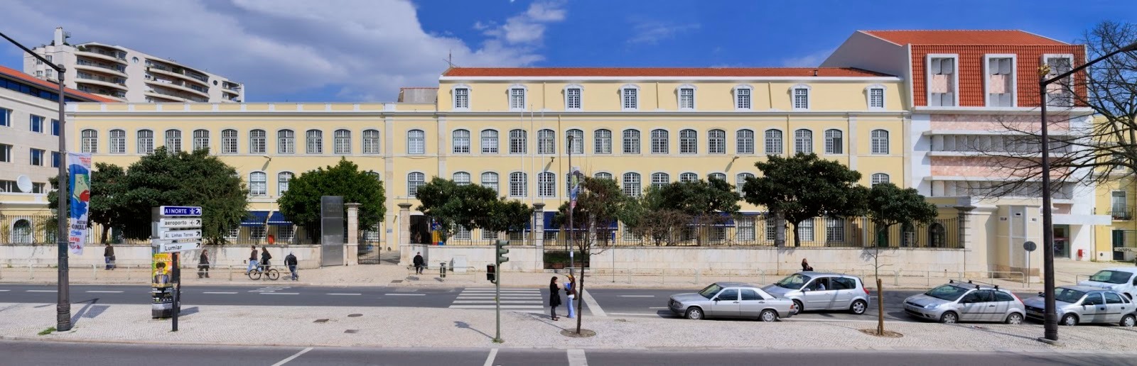 Panoramic street-level view of a long, pale yellow institutional building with three stories of evenly spaced arched windows and a red-tiled roof under a partly cloudy blue sky. A low stone wall and iron fence line the sidewalk in front of the building, with several large green trees breaking up the façade. In the foreground, cars are parked along the curb and a few pedestrians walk or stand near a crosswalk and traffic light. Directional road signs and a tall streetlight pole are visible on the left side of the image.