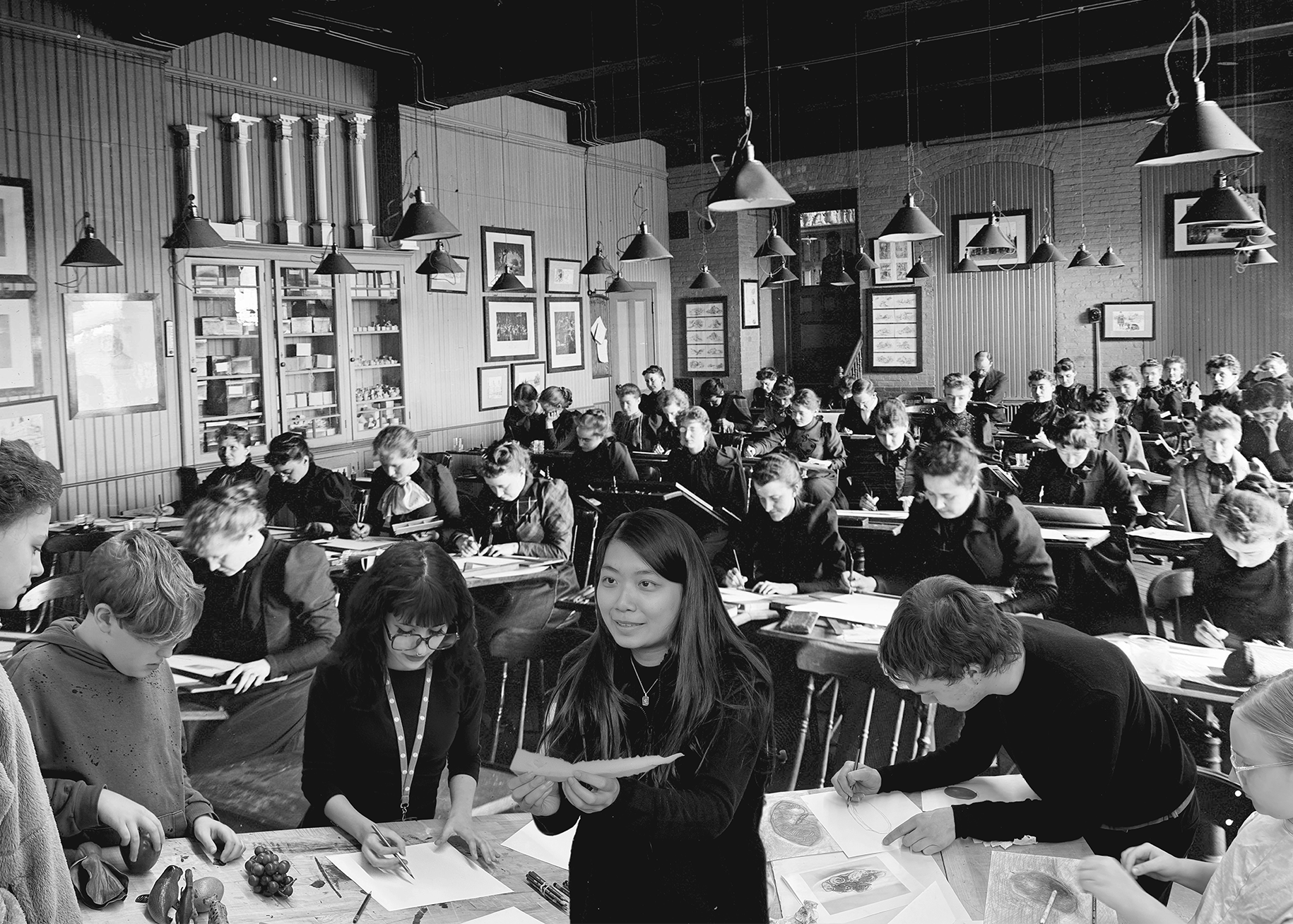 A black-and-white image of a classroom filled with students, predominantly boys, seated at desks focused on drawing or writing. In the foreground, a girl is holding a piece of paper and appears to be instructing a couple of peers. The classroom features high ceilings, hanging lights, and various educational materials displayed on the walls.