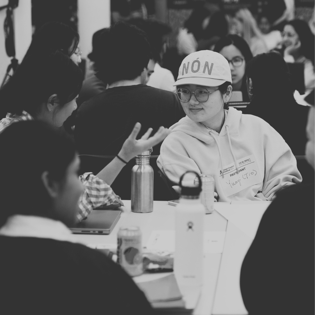 Black-and-white photograph of a crowded indoor student gathering. In the foreground, students sit around a table cluttered with water bottles, a laptop, and canned drinks. At the center, a student wearing glasses, a hoodie, and a baseball cap printed with the word “NON” listens attentively to another student gesturing mid-conversation. The background shows many additional students engaged in group discussions. The scene conveys an active, social, collaborative environment.
