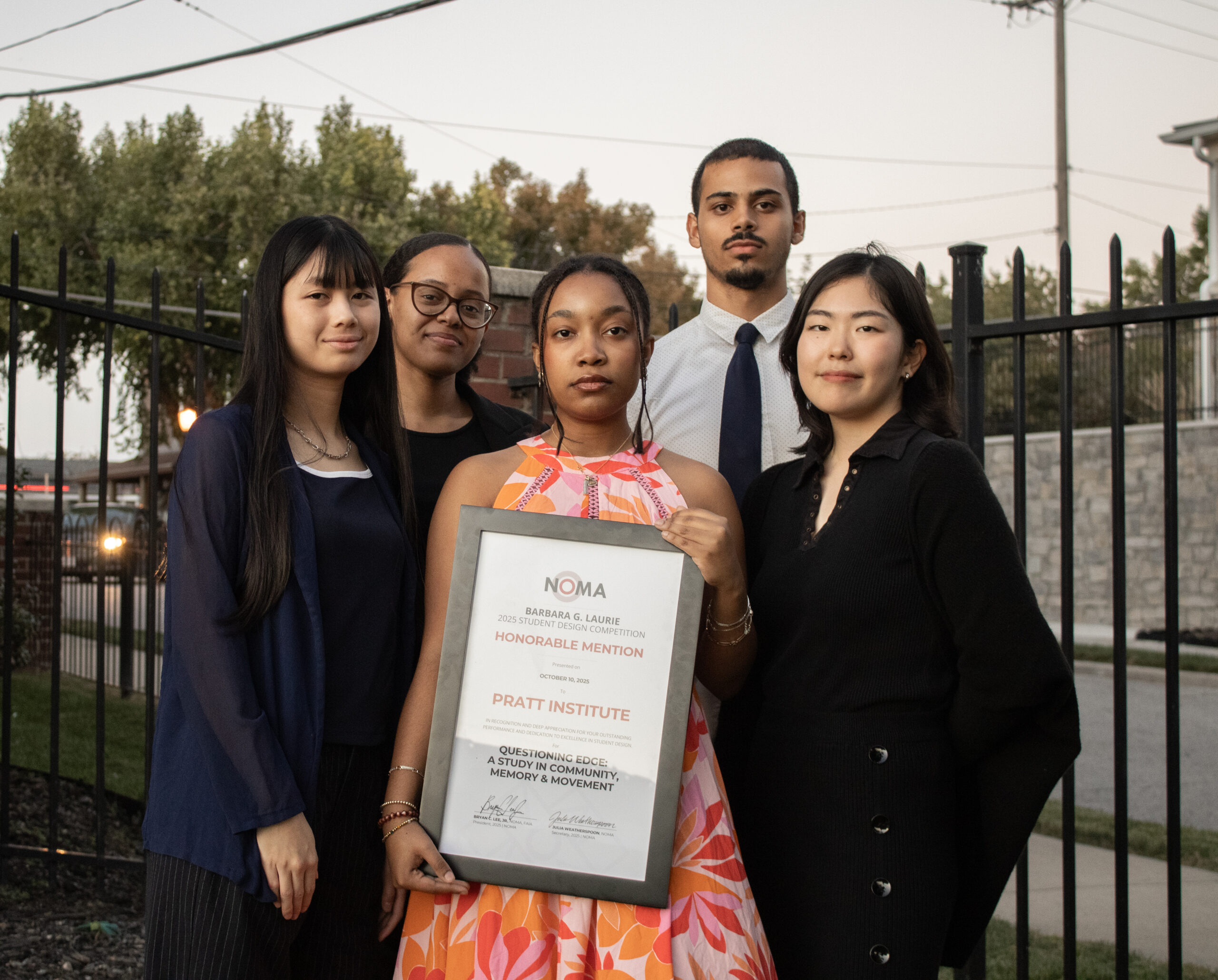 A group of five individuals stands together outdoors, smiling and posing for a photo. One person holds a certificate framed with the text "NOMA Barbara G. Laurie 2025 Student Design Competition Honorable Mention, Pratt Institute." They are dressed in a mix of casual and formal attire, with trees and a fence visible in the background, indicating it's late afternoon or early evening.