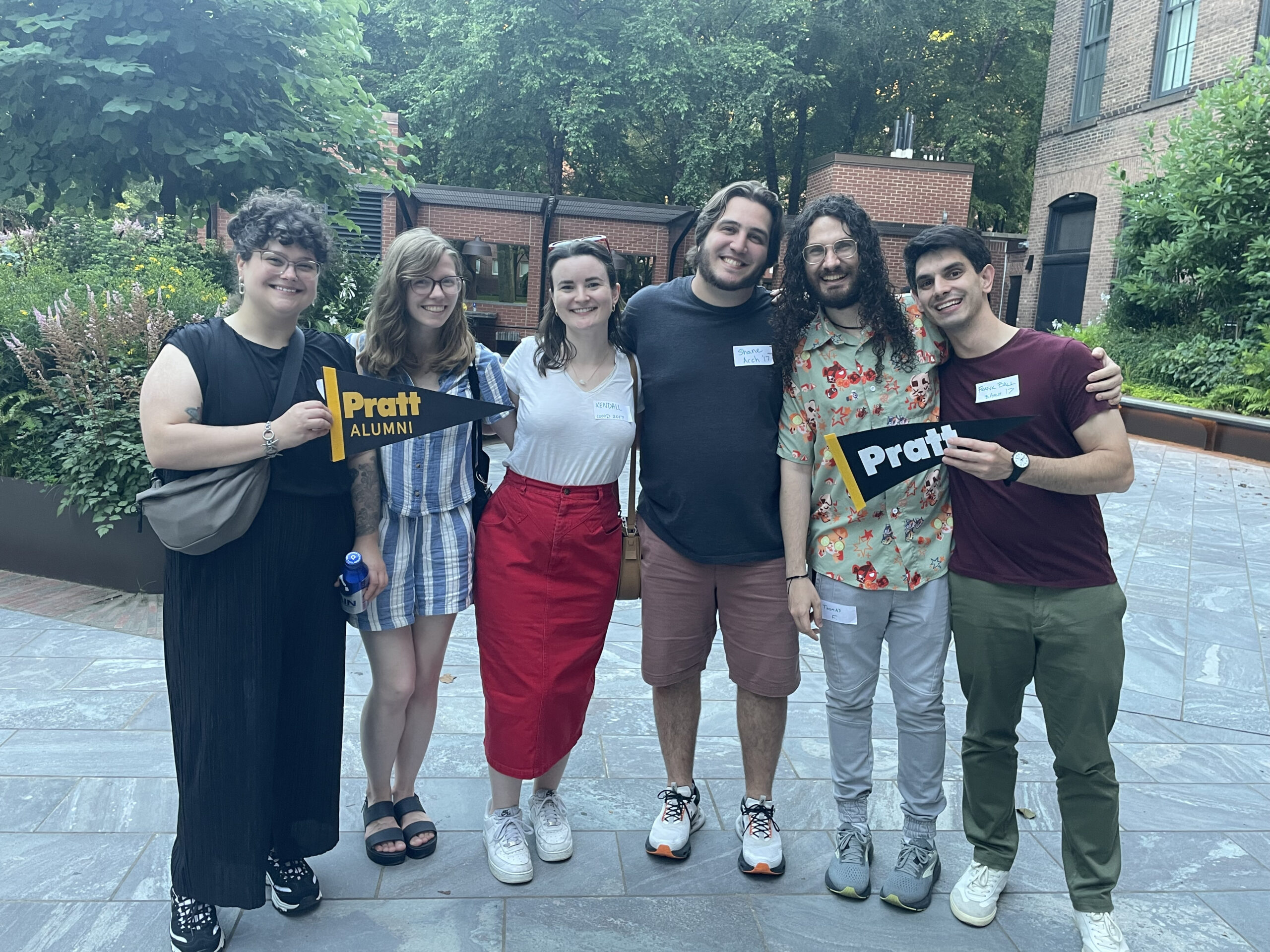 A group of six people stands together outdoors, smiling and posing for a photo. They are holding a black and yellow pennant that reads "Pratt ALUMNI." The background features greenery and brick buildings, indicating a social gathering. They are dressed casually, with a mix of shorts, dresses, and shirts.