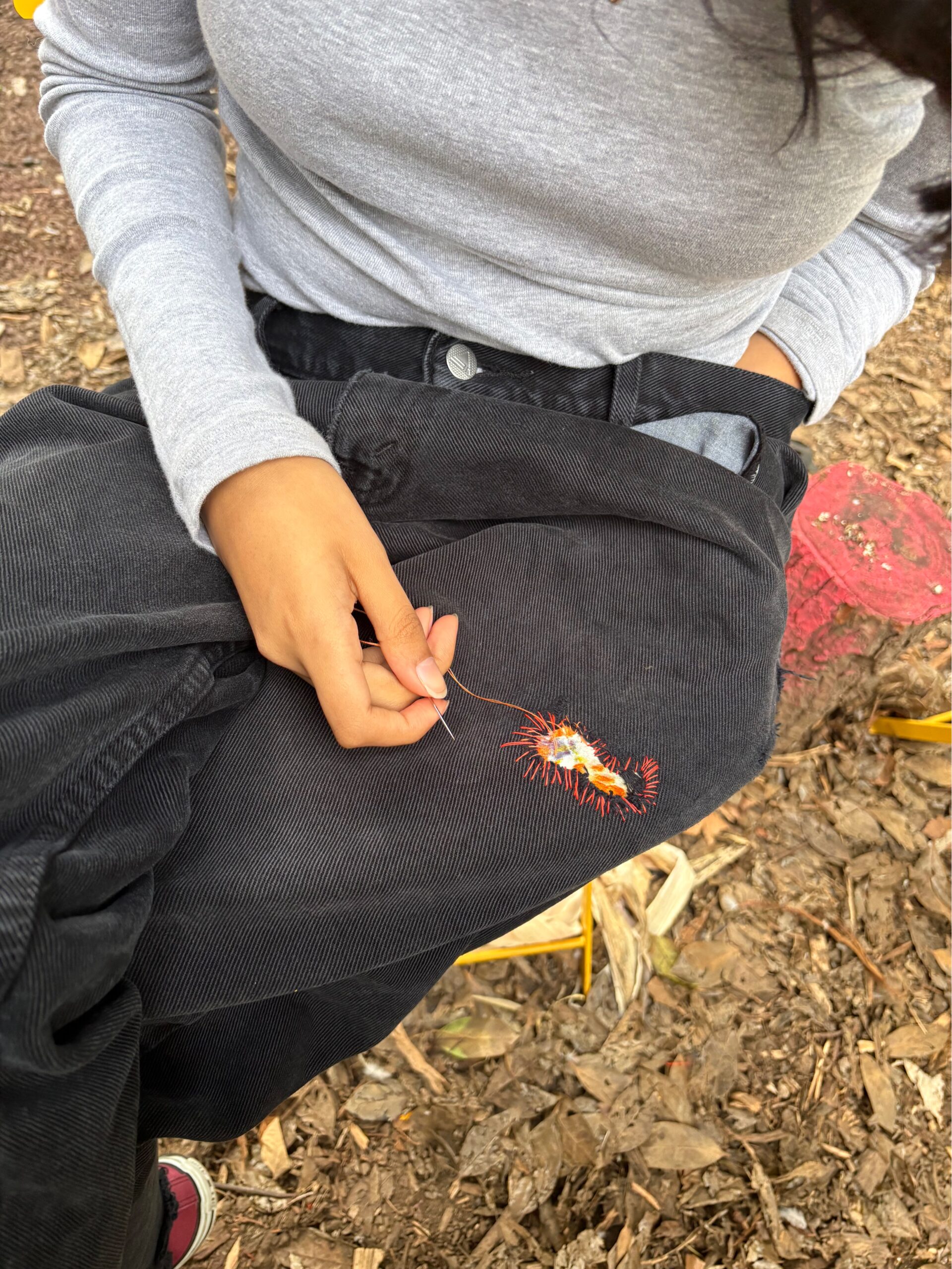 A close-up of a person sitting on the ground, wearing a light gray long-sleeve shirt and black corduroy pants. They are holding a needle and thread, working on an embroidered design featuring a flower with red threads. The ground is covered with leaves and there are hints of yellow objects in the background.