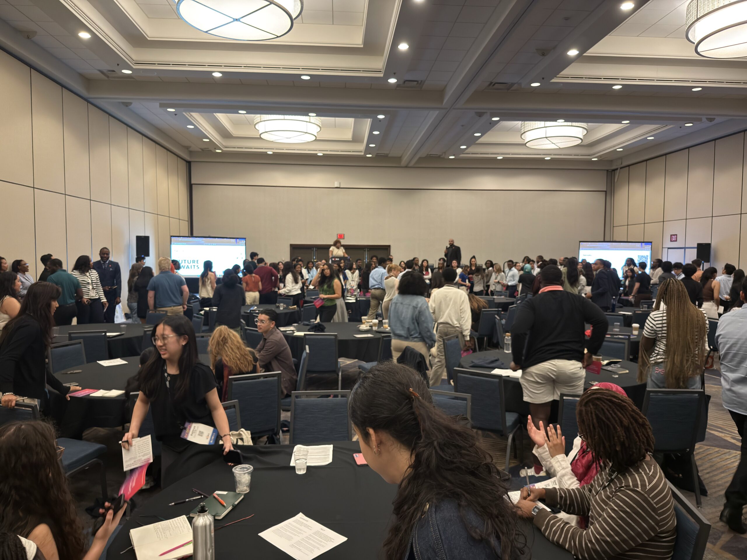 A large conference room filled with attendees interacting and networking. People are gathered around tables, some standing and others sitting, with screens visible in the background displaying information. The atmosphere is lively and collaborative, showcasing a diverse group of individuals engaged in conversation.