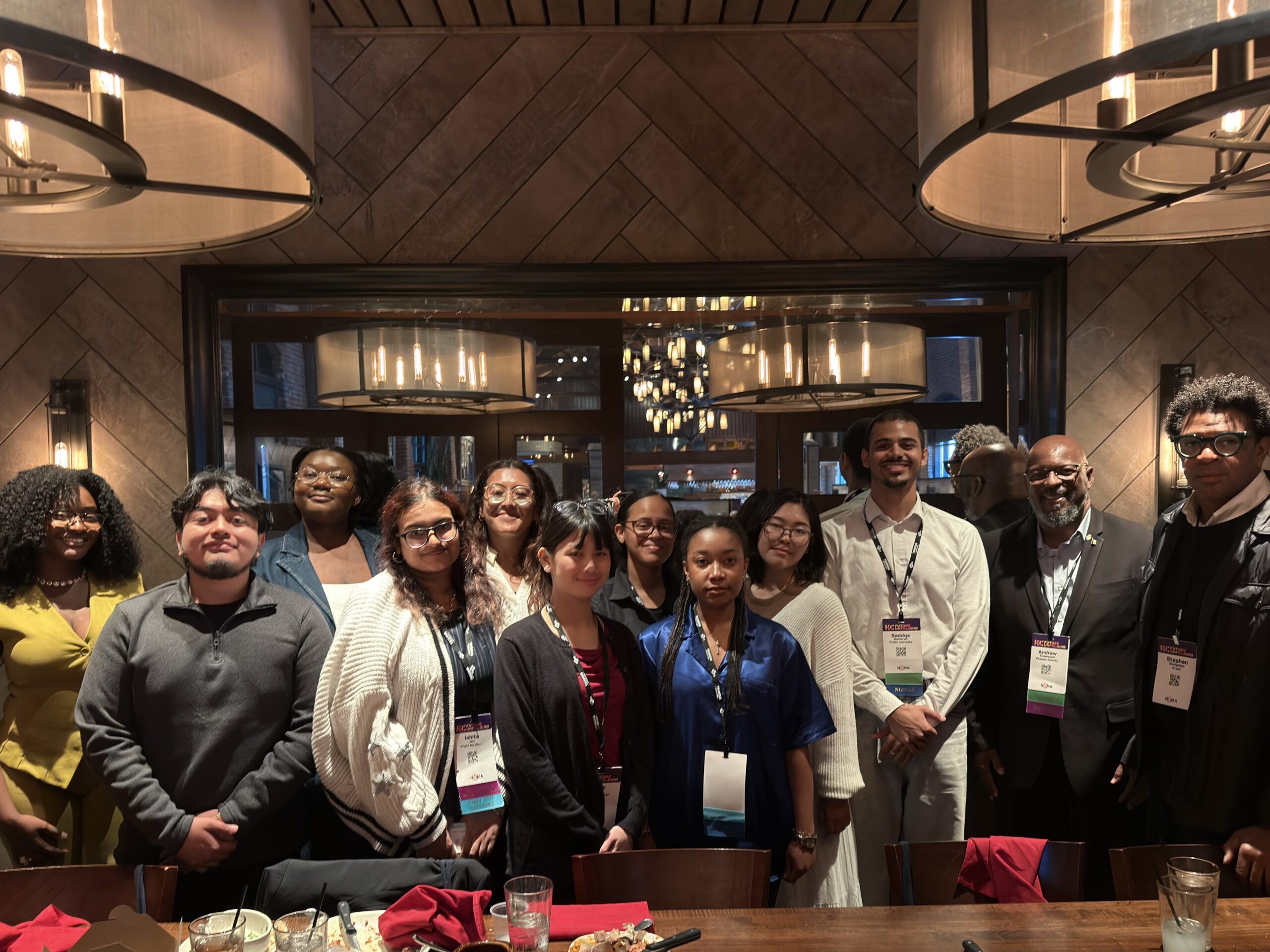 A large conference room filled with attendees interacting and networking. People are gathered around tables, some standing and others sitting, with screens visible in the background displaying information. The atmosphere is lively and collaborative, showcasing a diverse group of individuals engaged in conversation.